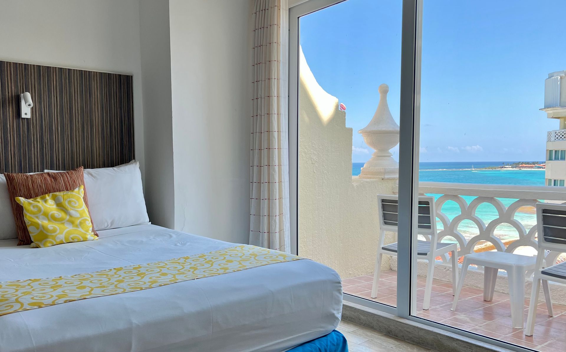 Dormitorio con vista al mar, cama con detalles amarillos, agua azul, día soleado.