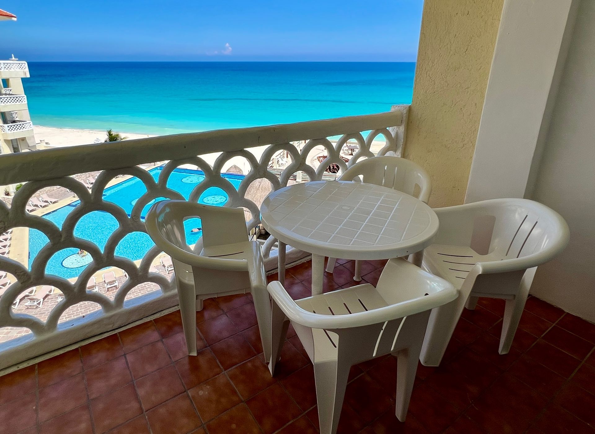 Balcón con sillas blancas y mesa con vista a la piscina y al mar turquesa.