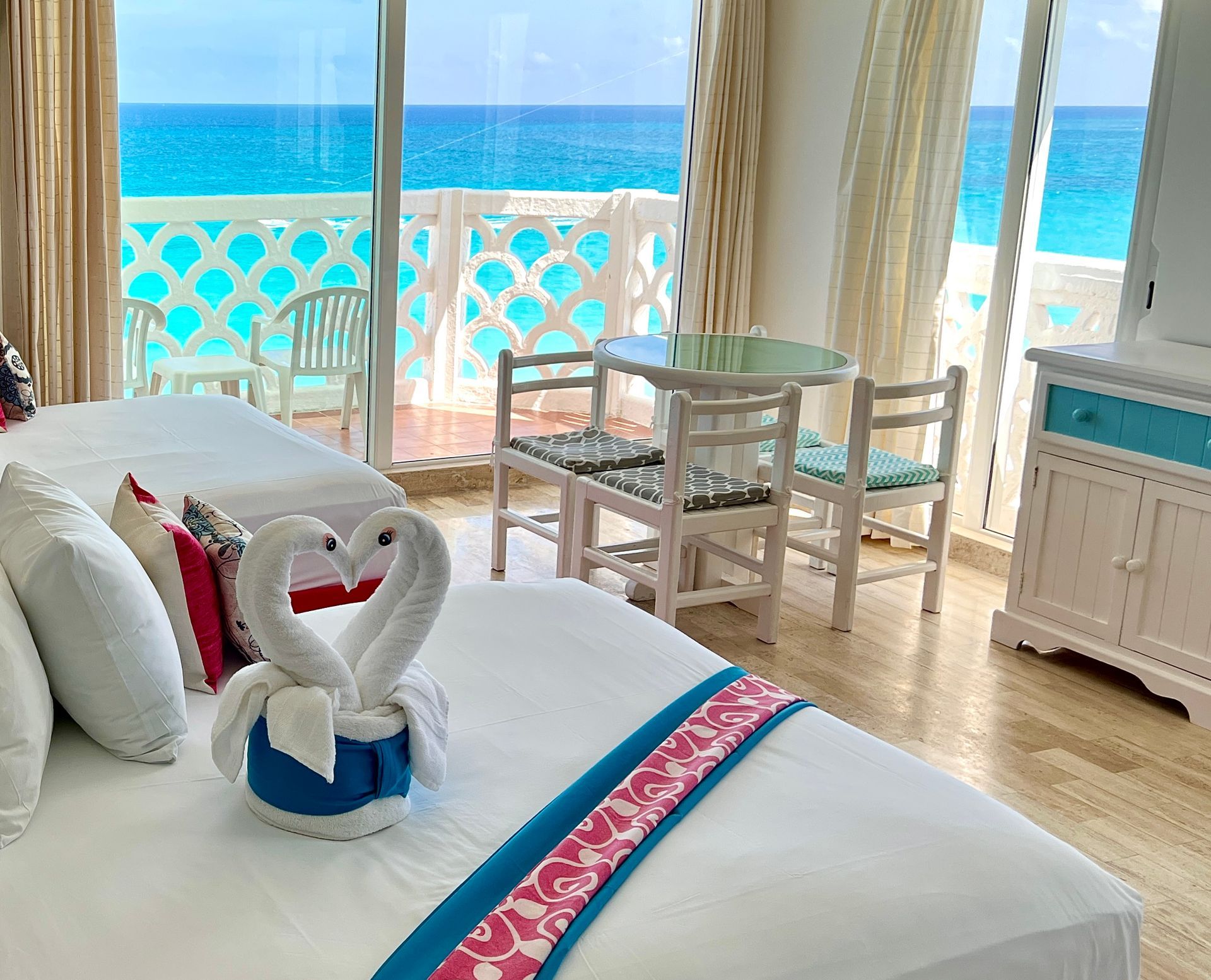 Bright hotel room with ocean view, white beds, teal accents, small table, balcony.