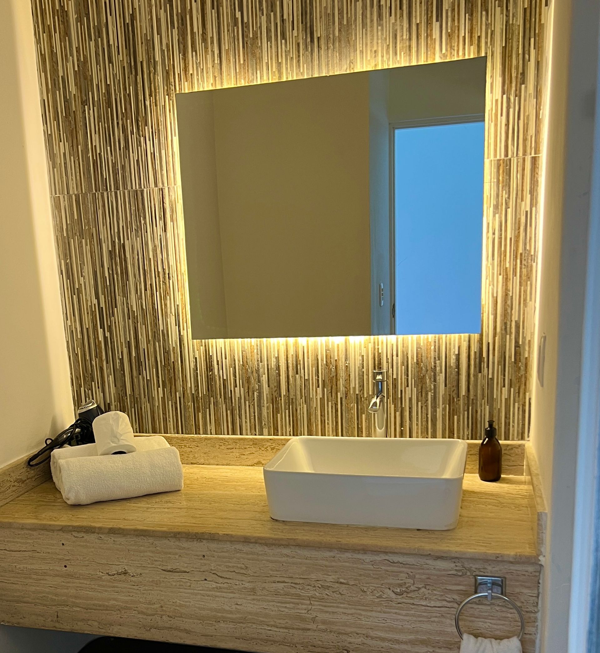 Baño con espejo iluminado, lavabo blanco, encimera de piedra y pared estampada.