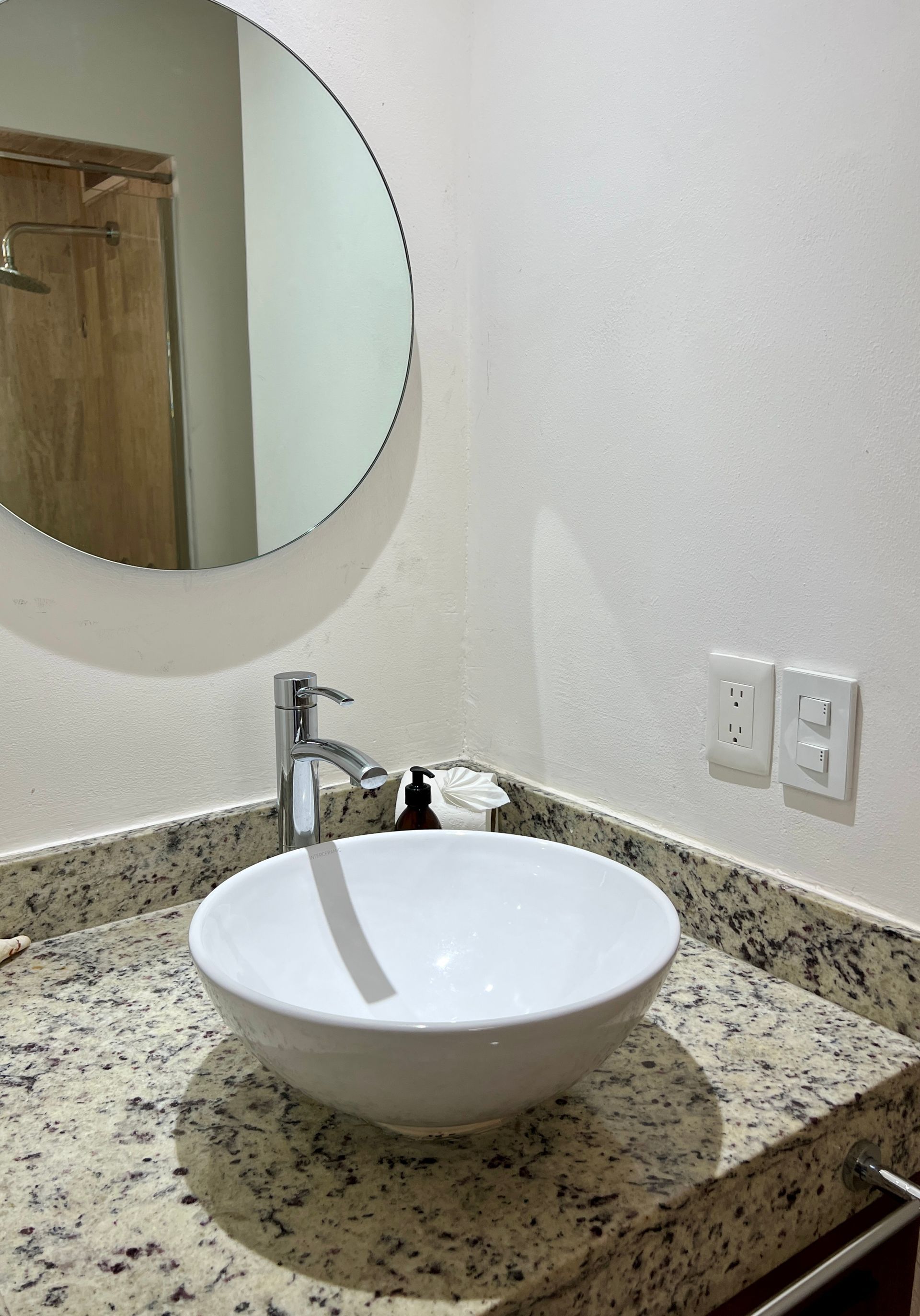 Mueble de baño con lavabo redondo, espejo, encimera de granito y tomas de pared.
