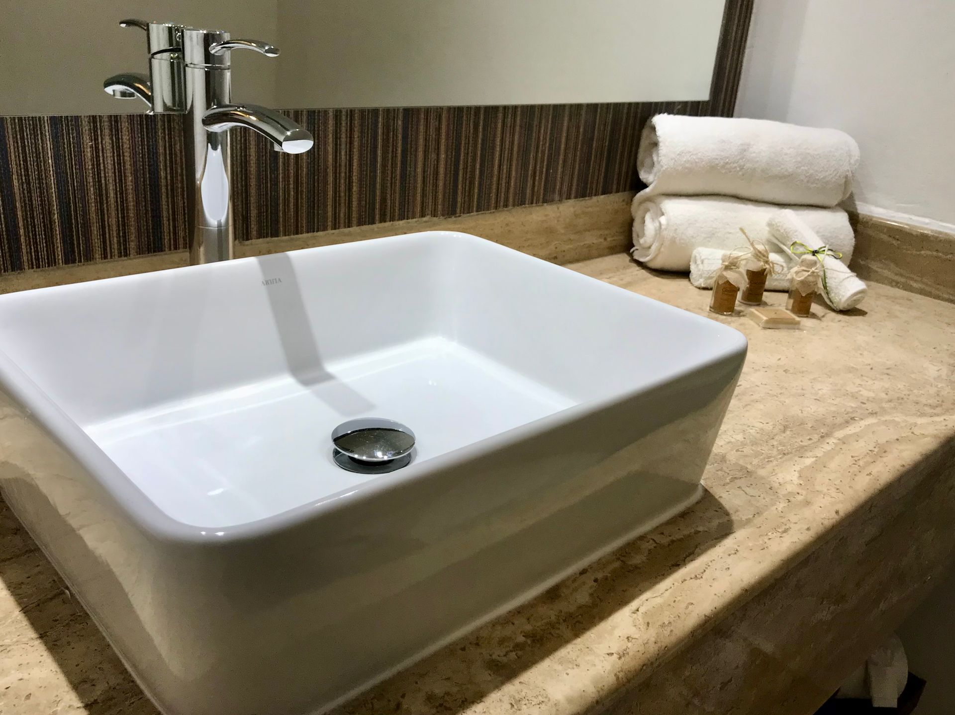 Lavabo rectangular blanco con grifería cromada sobre encimera de mármol, toallas y artículos de aseo al fondo.