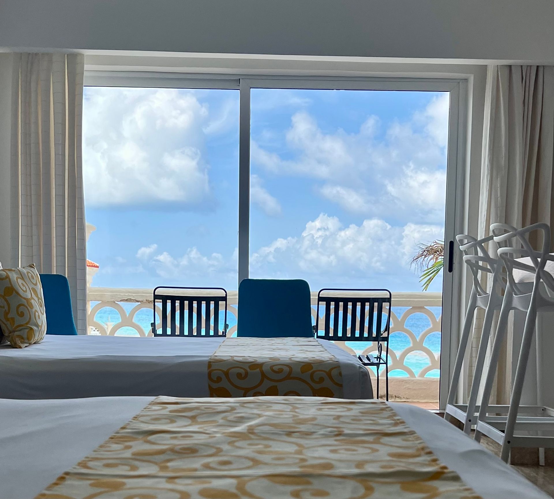 Dormitorio con vista al mar. Dos camas, balcón con sillas azules, cortinas y un cielo radiante.