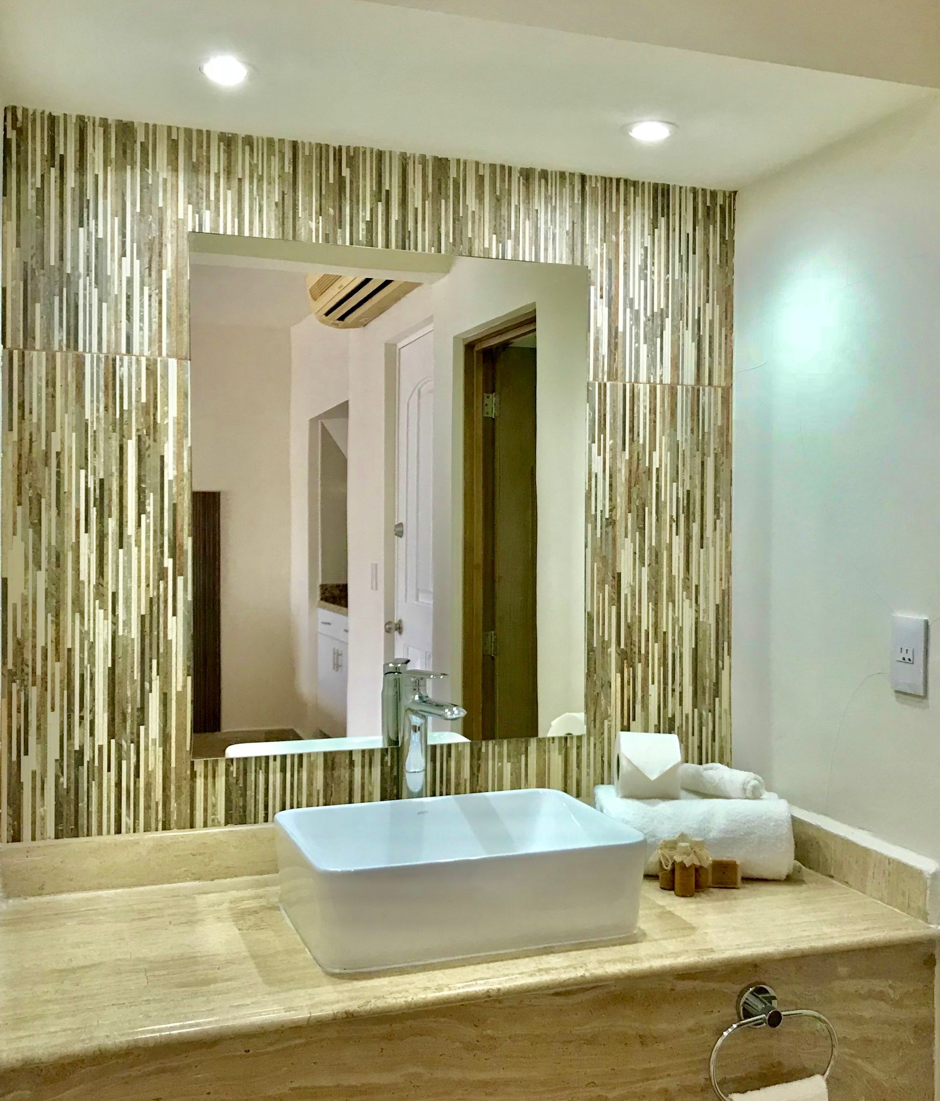 Tocador de baño con espejo cuadrado rodeado de azulejos texturizados, lavabo blanco y toallas.