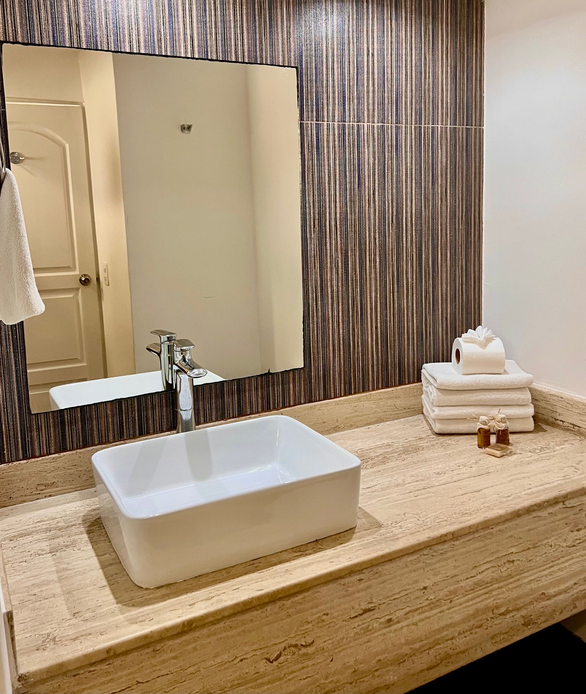 Baño con lavabo rectangular blanco sobre encimera beige, espejo y pared marrón texturizada.
