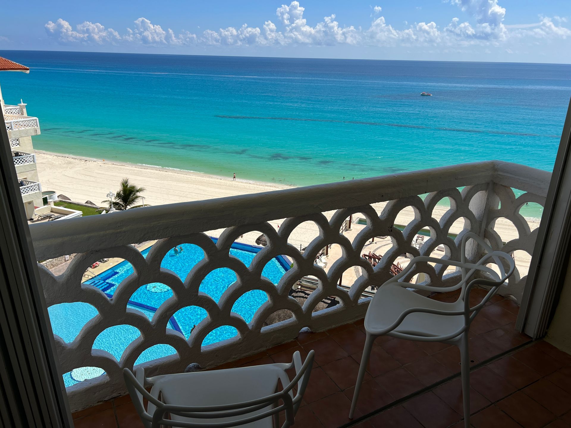 Balcón con vista al mar: agua turquesa, playa de arena blanca, piscina, sillas blancas, cielo azul.