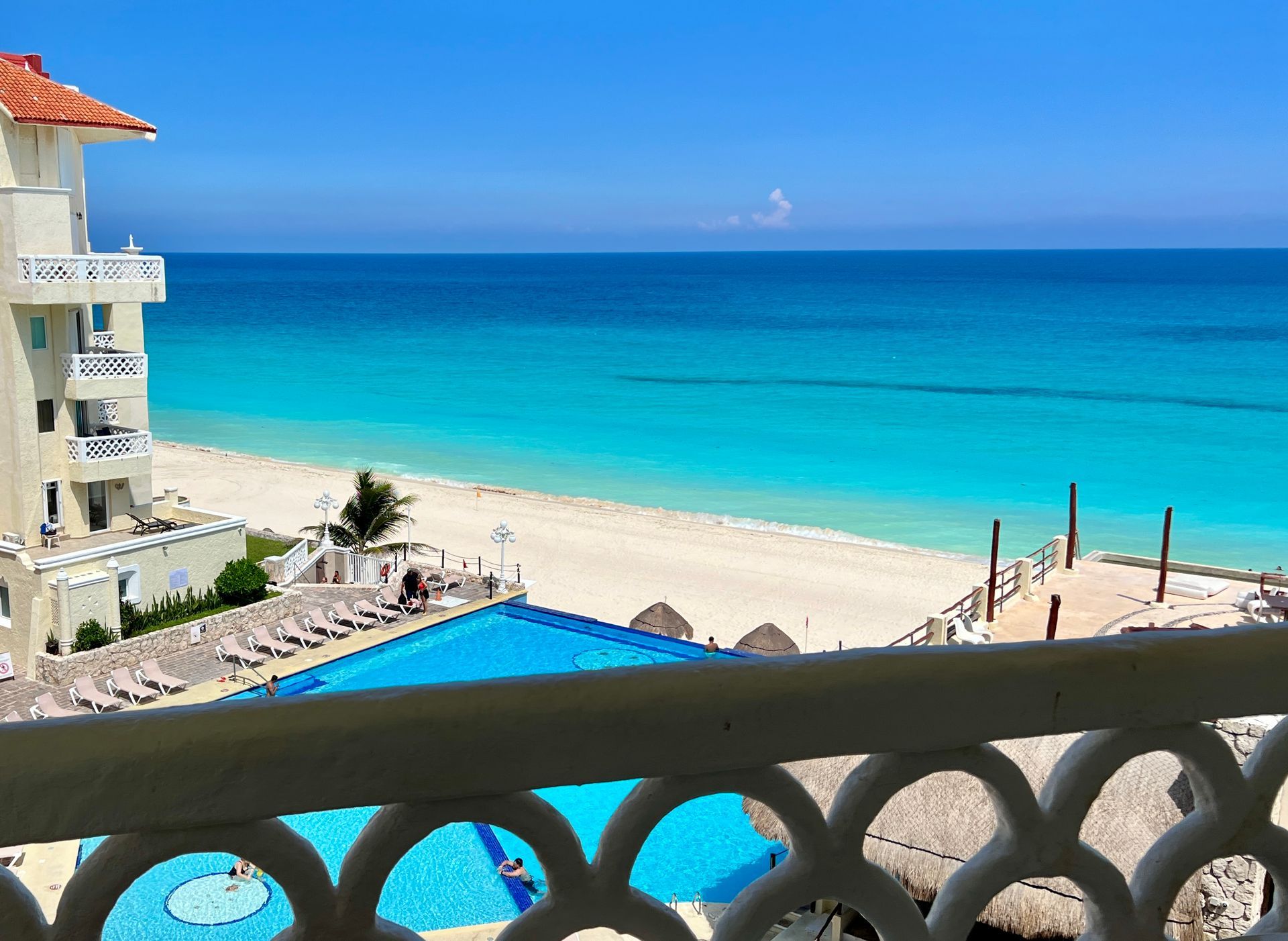 Vista desde un balcón con vista a un océano turquesa, playa de arena blanca, piscina y edificio del resort.