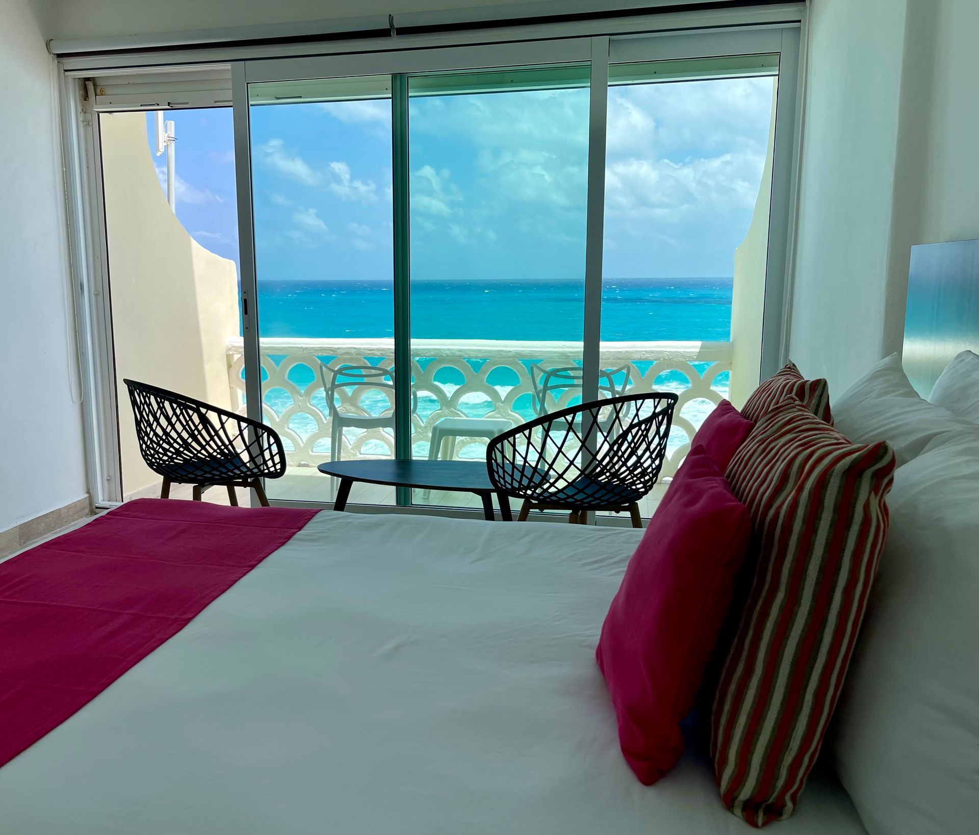Dormitorio con vista al mar, cama con almohadas y manta rosa y roja, dos sillas negras y mesa pequeña en balcón.