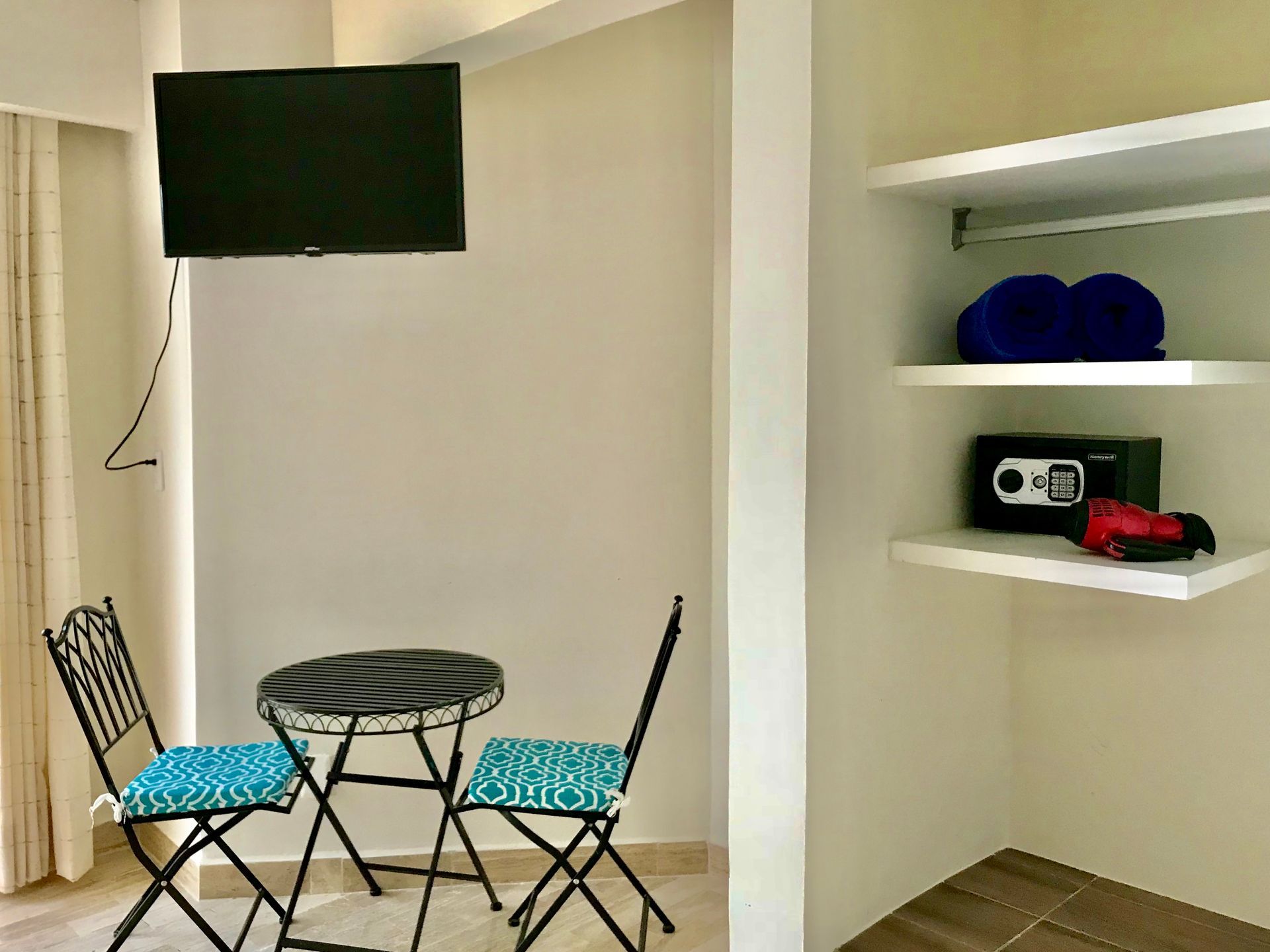 Habitación con mesa pequeña y sillas, TV y estanterías empotradas con toallas, caja fuerte y secador de pelo.