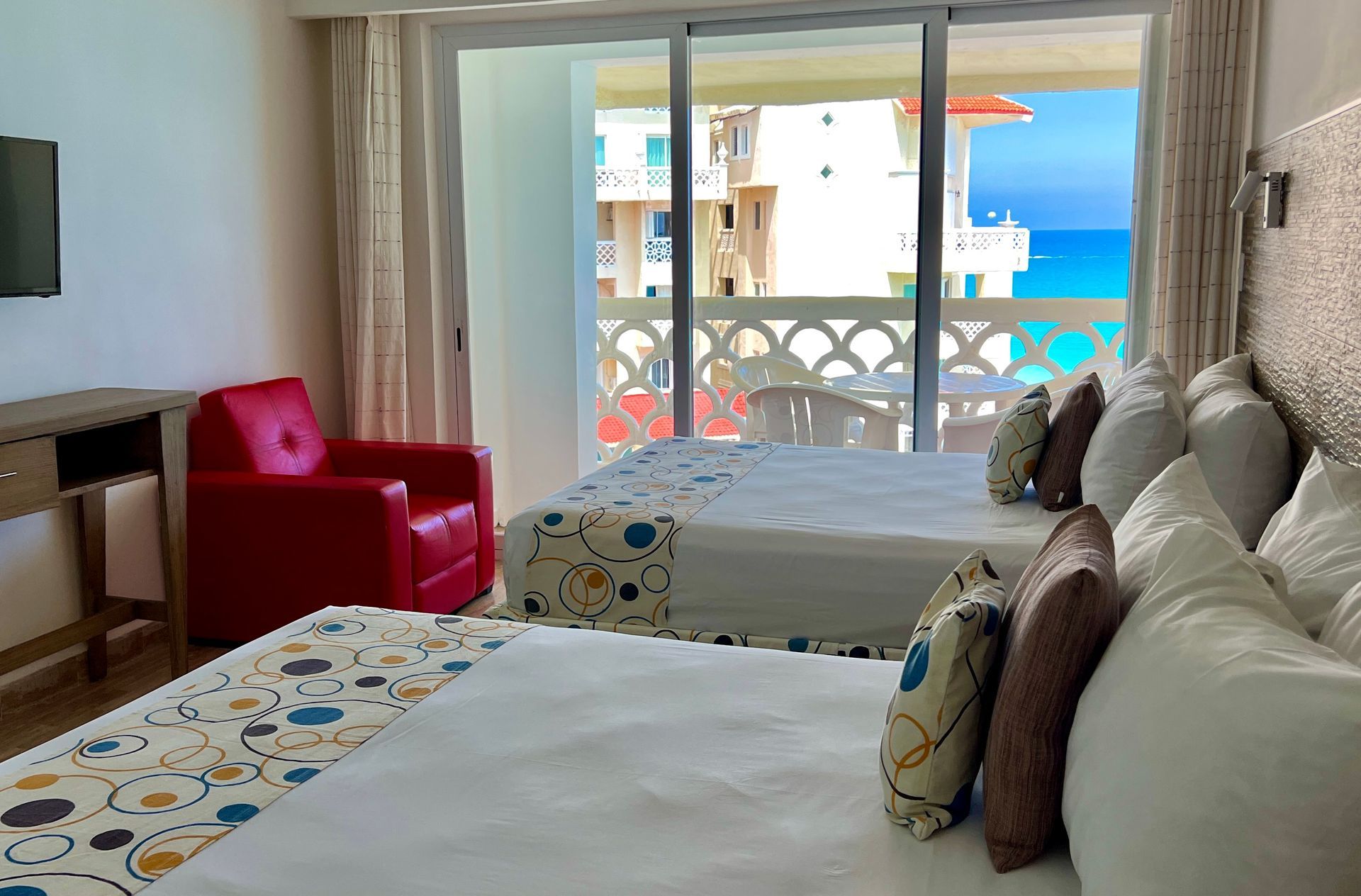 Dos camas en habitación de hotel con vista al mar, silla roja, balcón y tv.