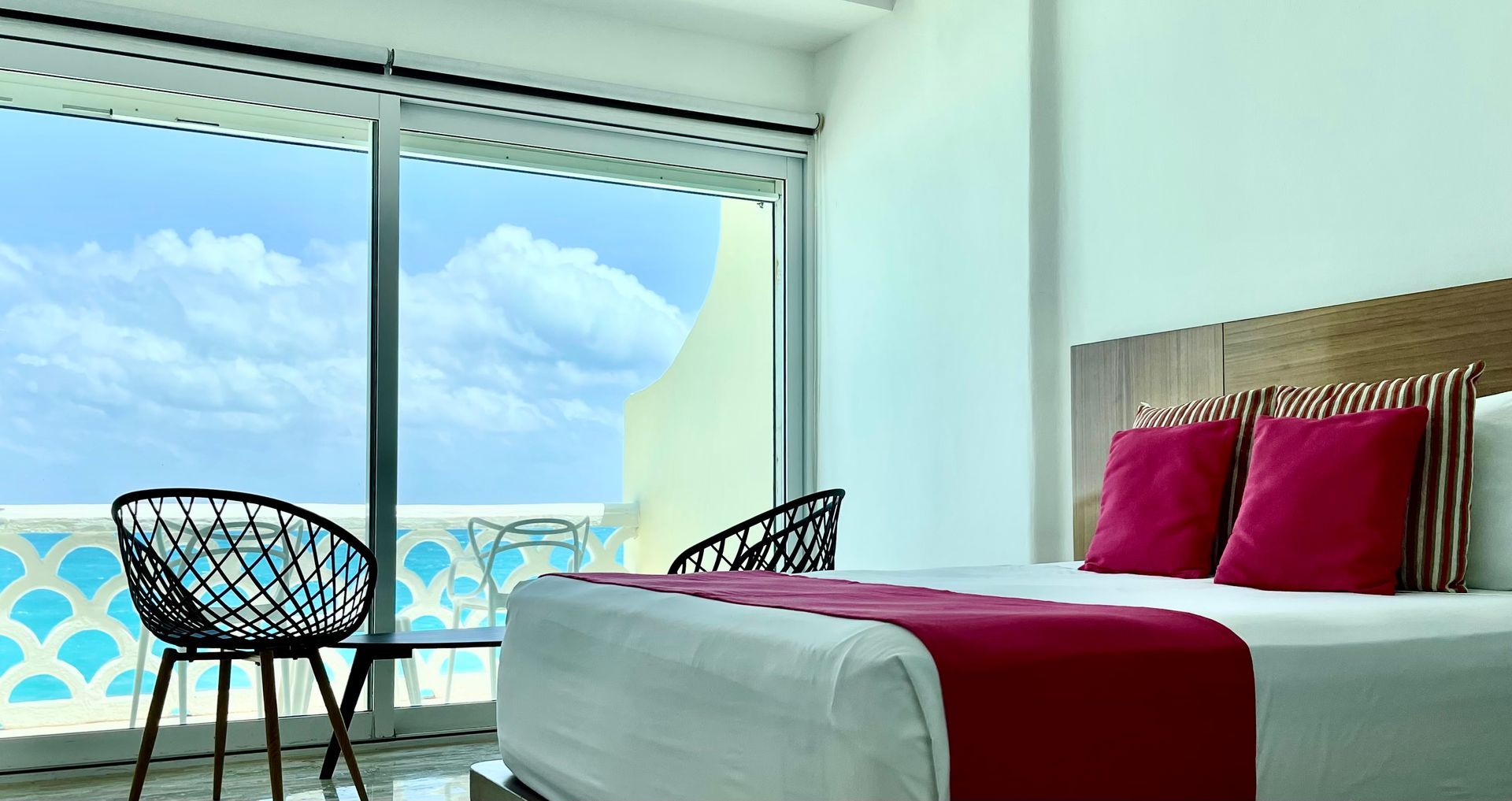 Habitación de hotel con cama, balcón, sillas y vista al mar y al cielo.