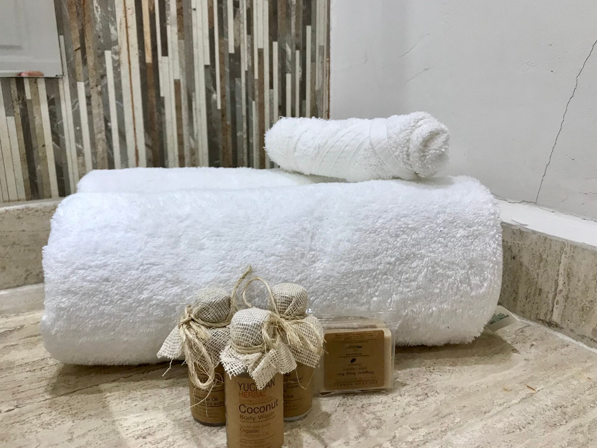 Toallas blancas y artículos de tocador sobre un mostrador de baño con protector contra salpicaduras de azulejos texturizados.