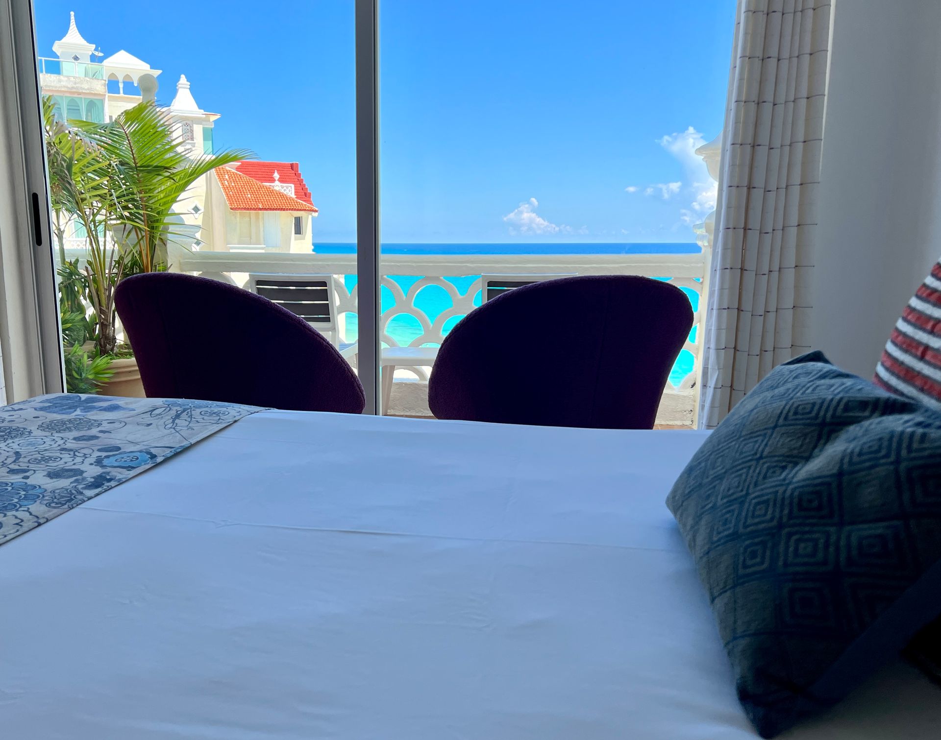 Dormitorio con vista al mar, dos sillas color morado oscuro, cama blanca, cielo azul.