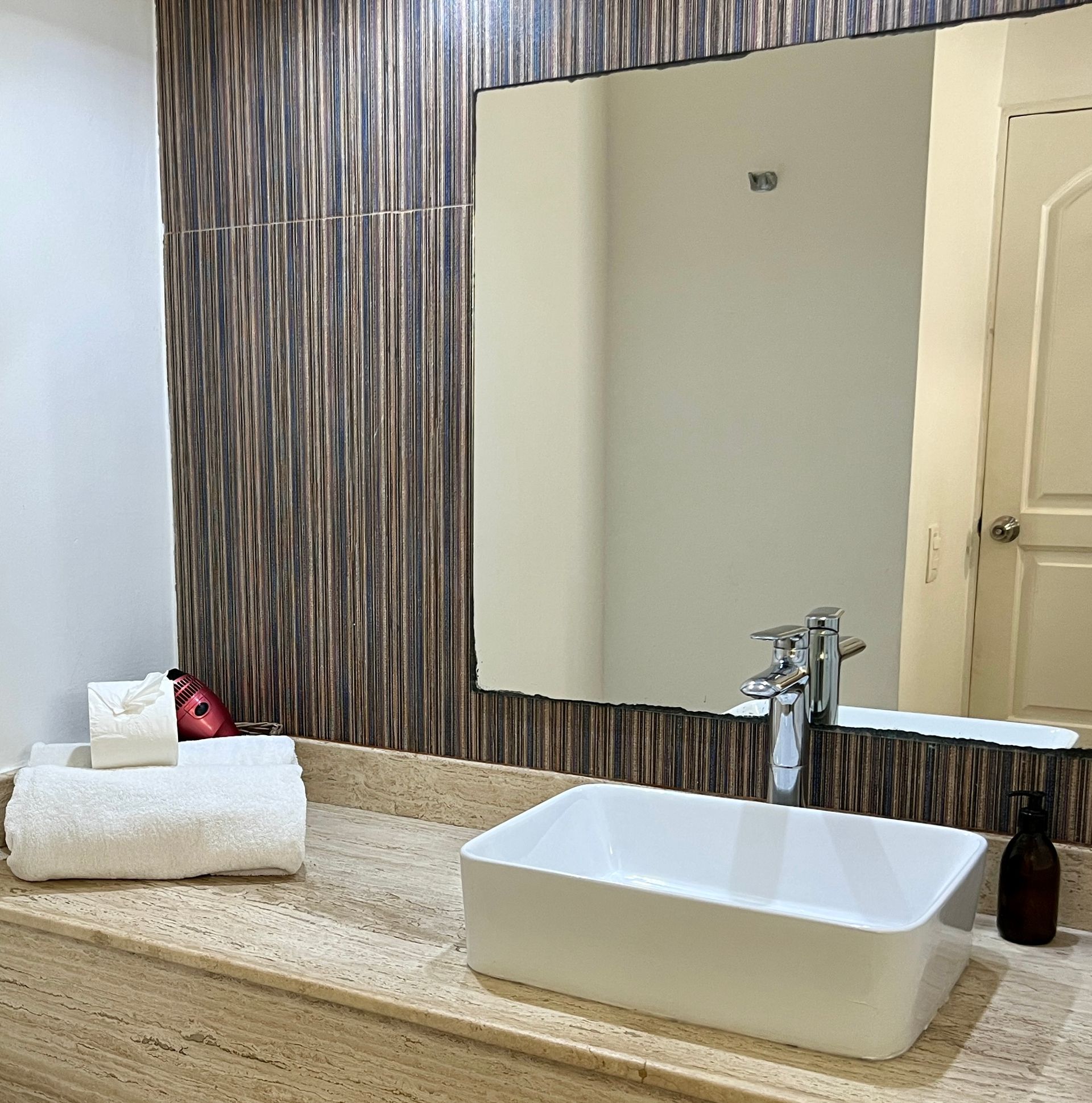 Baño moderno con lavabo blanco, espejo y pared con azulejos marrones. Toallas blancas en la encimera.