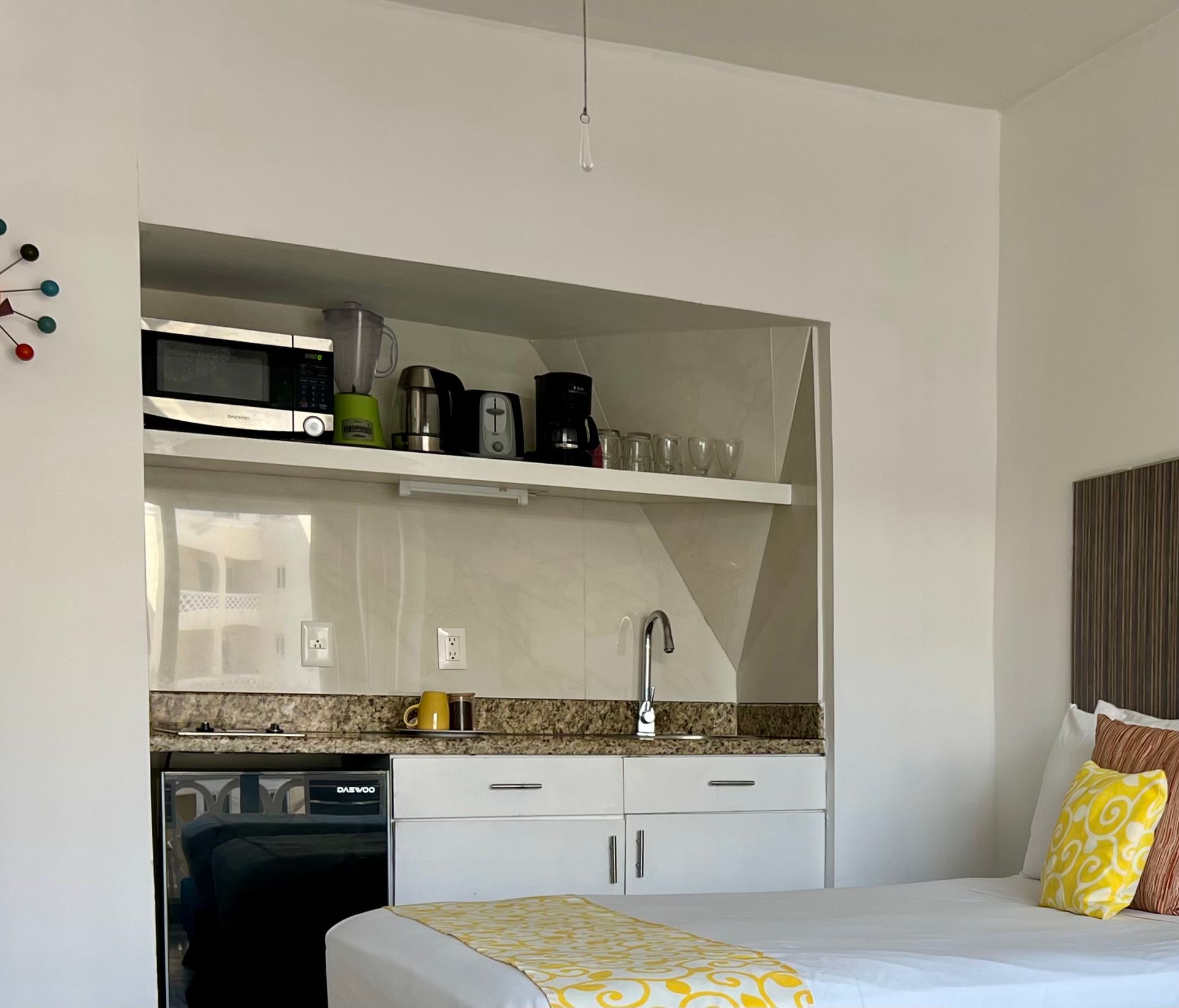Cocina tipo estudio con microondas, cafetera y fregadero. Cama con detalles en amarillo.