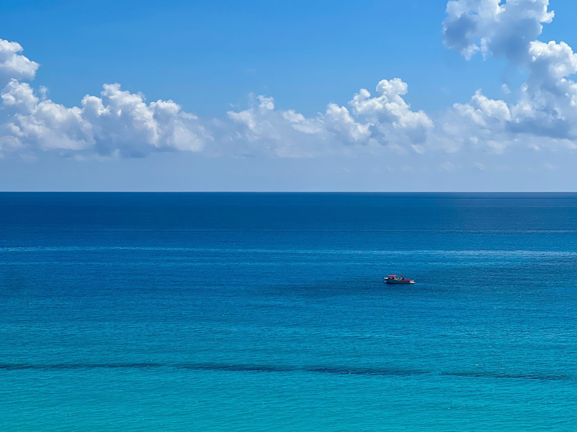 Océano azul vibrante bajo un cielo azul brillante con nubes blancas esponjosas; un pequeño bote flota.