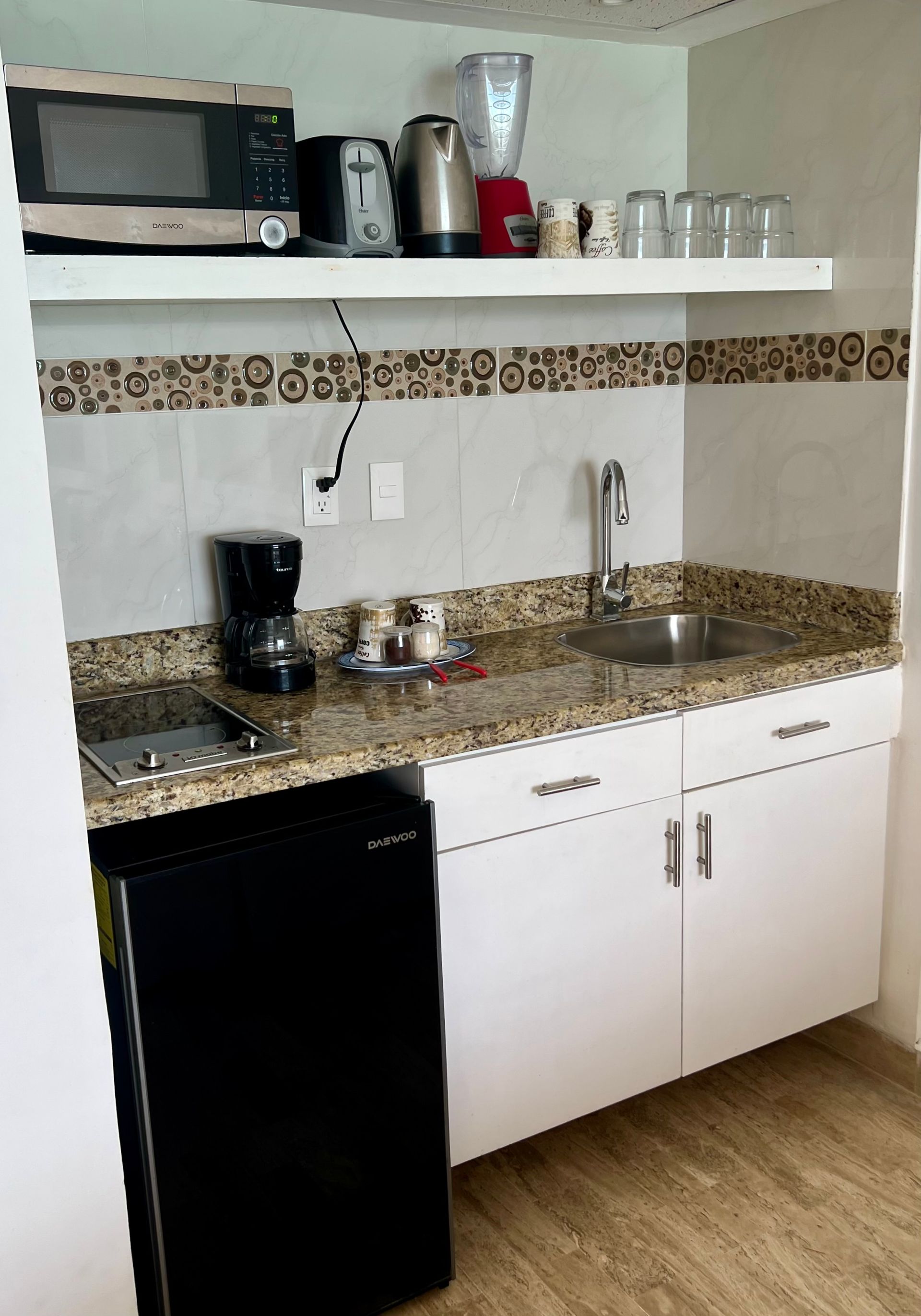 Pequeña cocina con refrigerador, microondas, cafetera, fregadero, gabinetes blancos y encimeras de granito.
