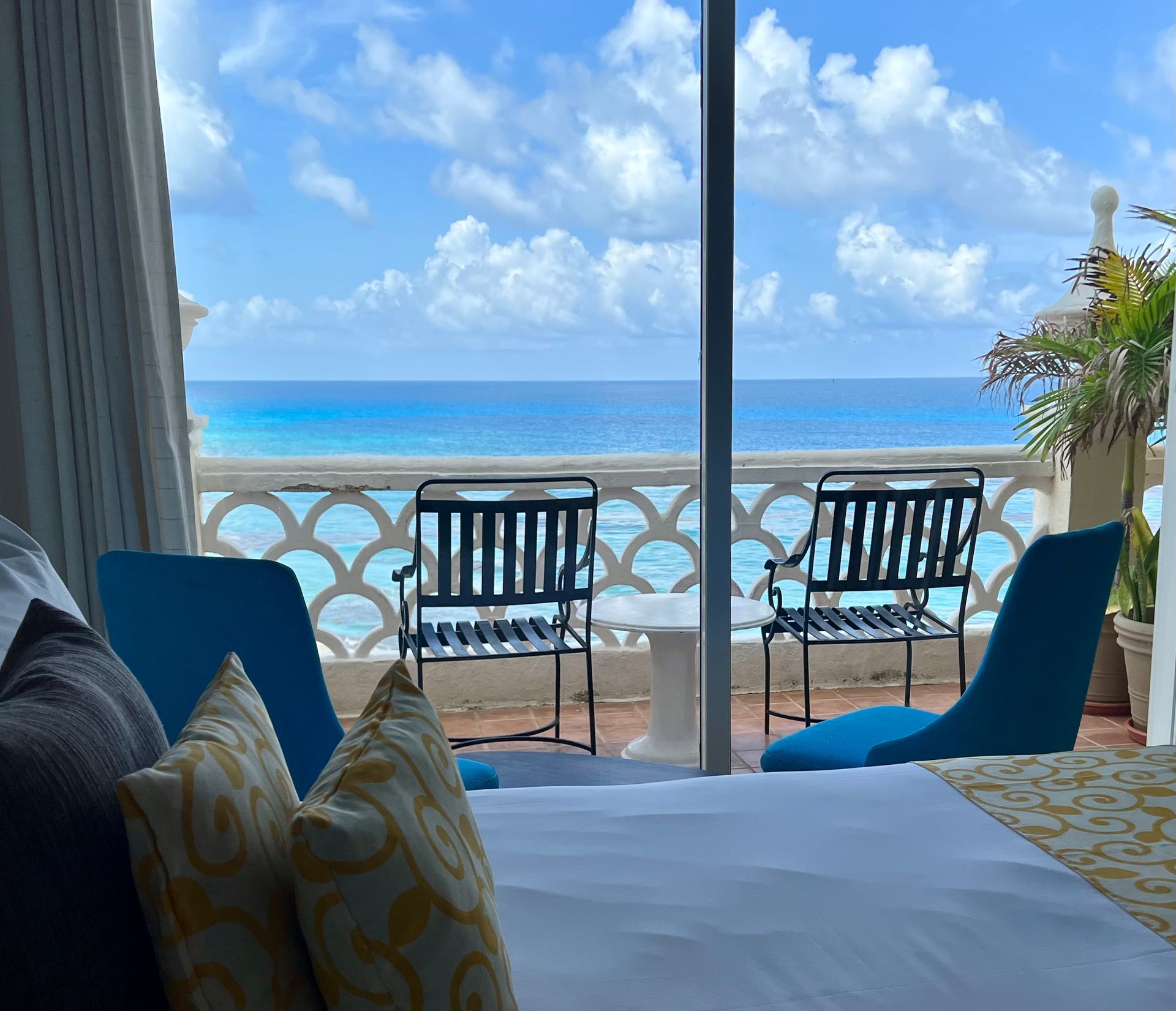 Bedroom with a balcony overlooking the turquoise ocean and blue sky. Two chairs and a small table on the balcony.