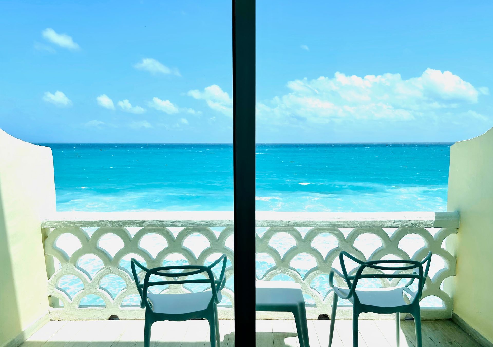 Balcón con vista al mar turquesa, dos sillas, barandilla decorativa blanca, cielo azul.