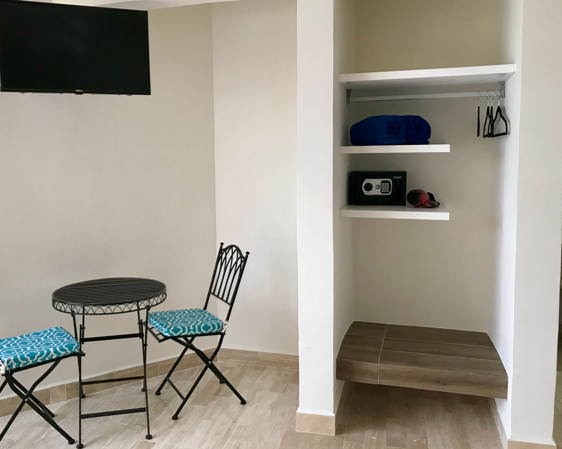 Una pequeña habitación de hotel con una mesa y sillas, estantes con artículos y un televisor en la pared.