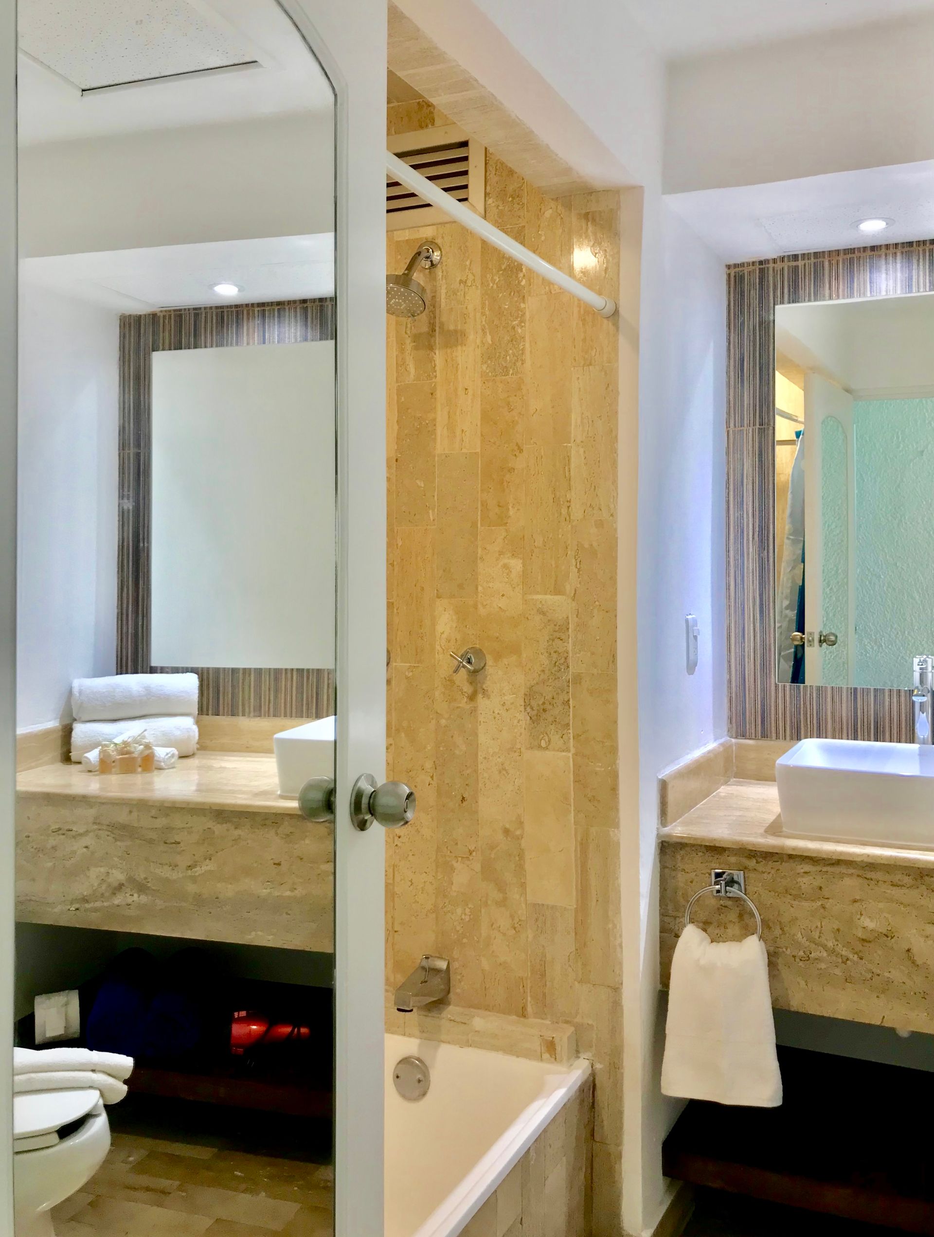 Baño con azulejos de piedra de color claro, lavabo y bañera blancos, espejo grande y zona de ducha con puerta de cristal.