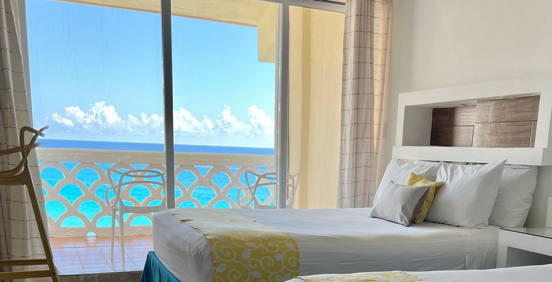 Dormitorio con dos camas con vista a balcón y vista al mar; sábanas blancas y amarillas.