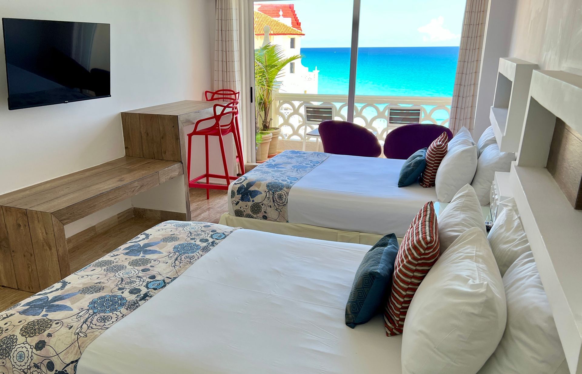 Habitación con dos camas y balcón con vista al mar. Taburetes rojos y televisor.