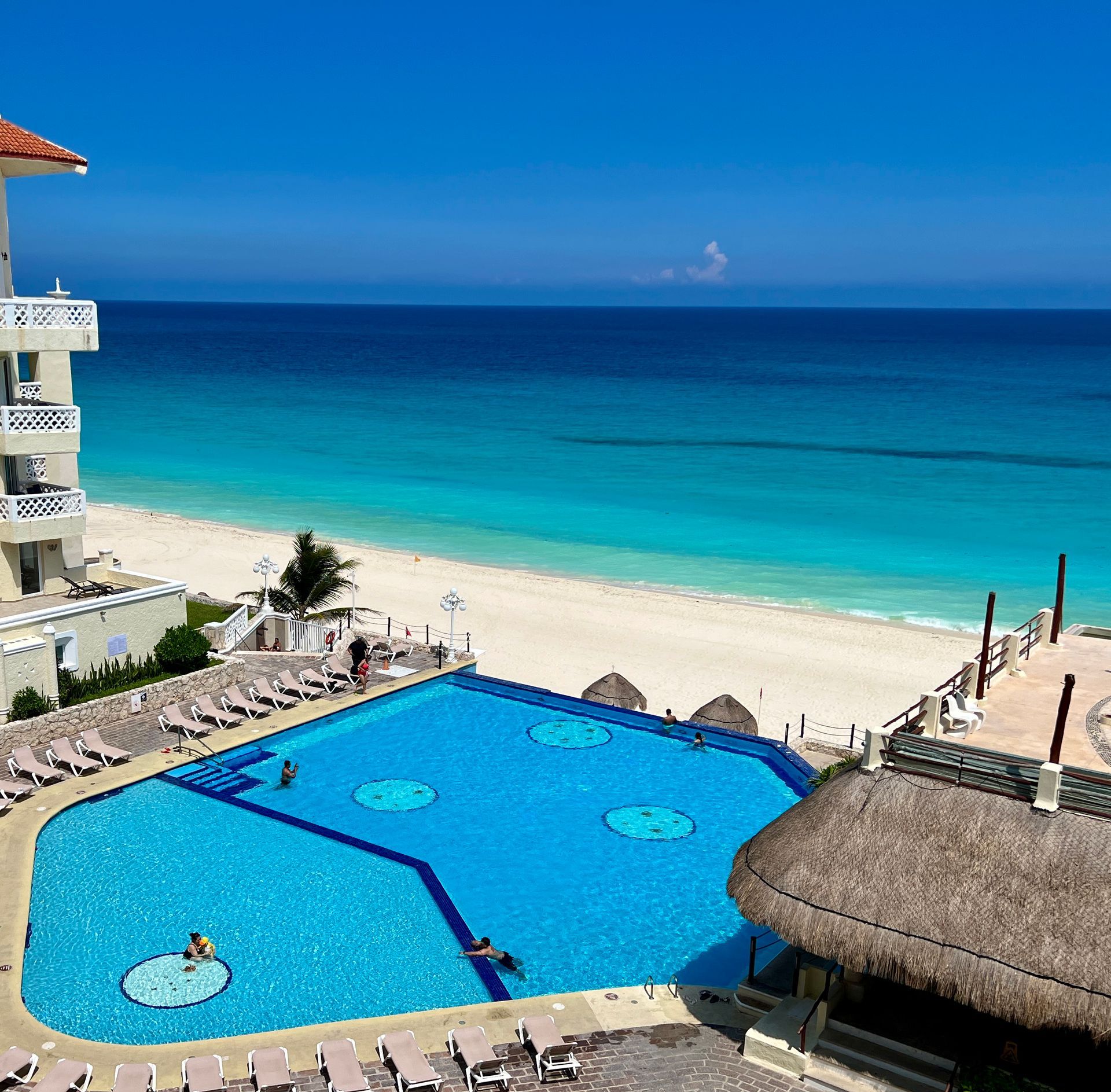 Vista de piscina y playa: agua turquesa, arena blanca, cielo azul, edificio a la izquierda y gente disfrutando.
