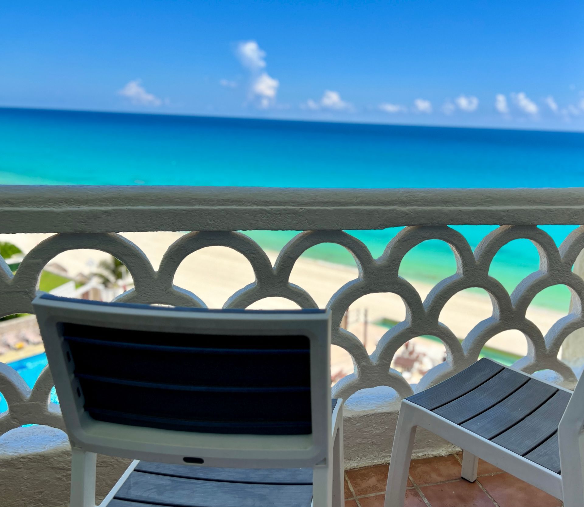Vista desde el balcón al océano y la playa, con barandilla decorativa, dos sillas, cielo azul y agua turquesa.