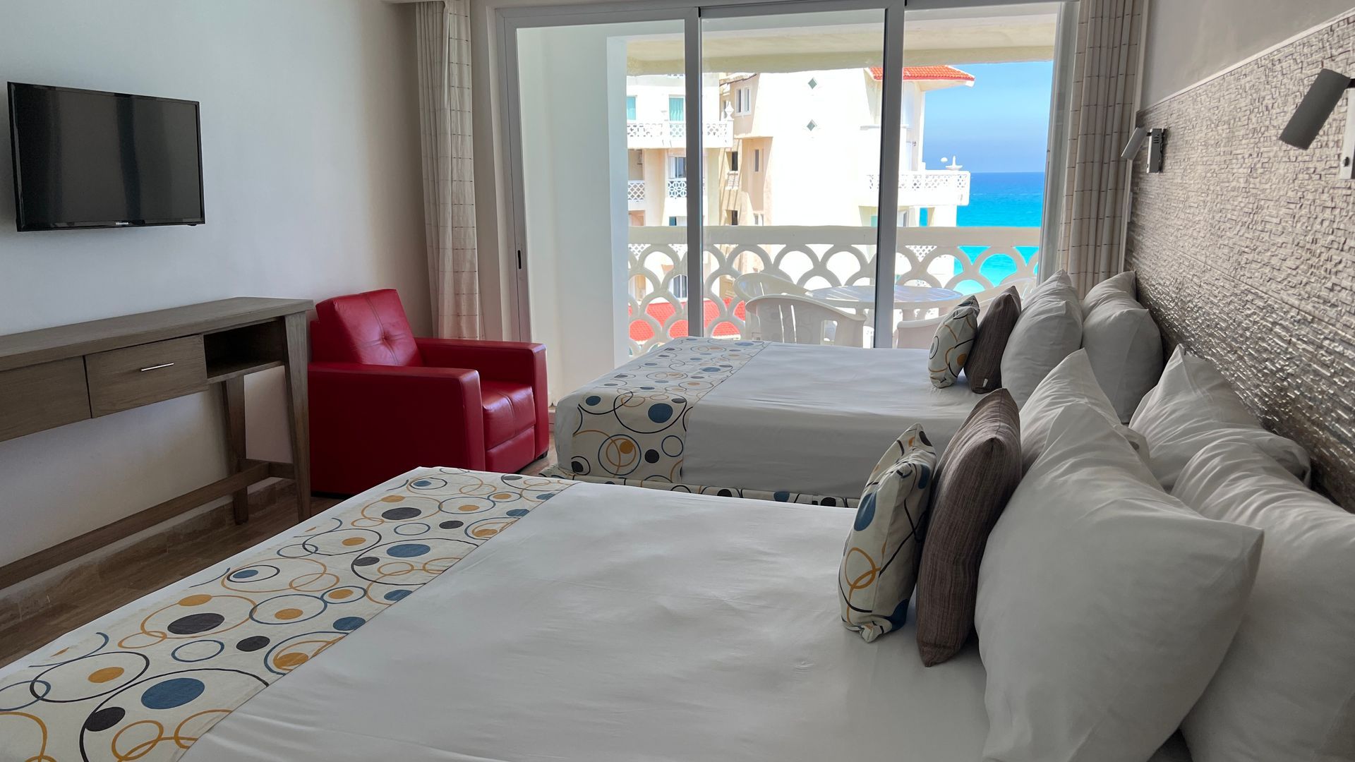 Habitación de hotel con dos camas, vista al mar, sillón rojo, TV y balcón.