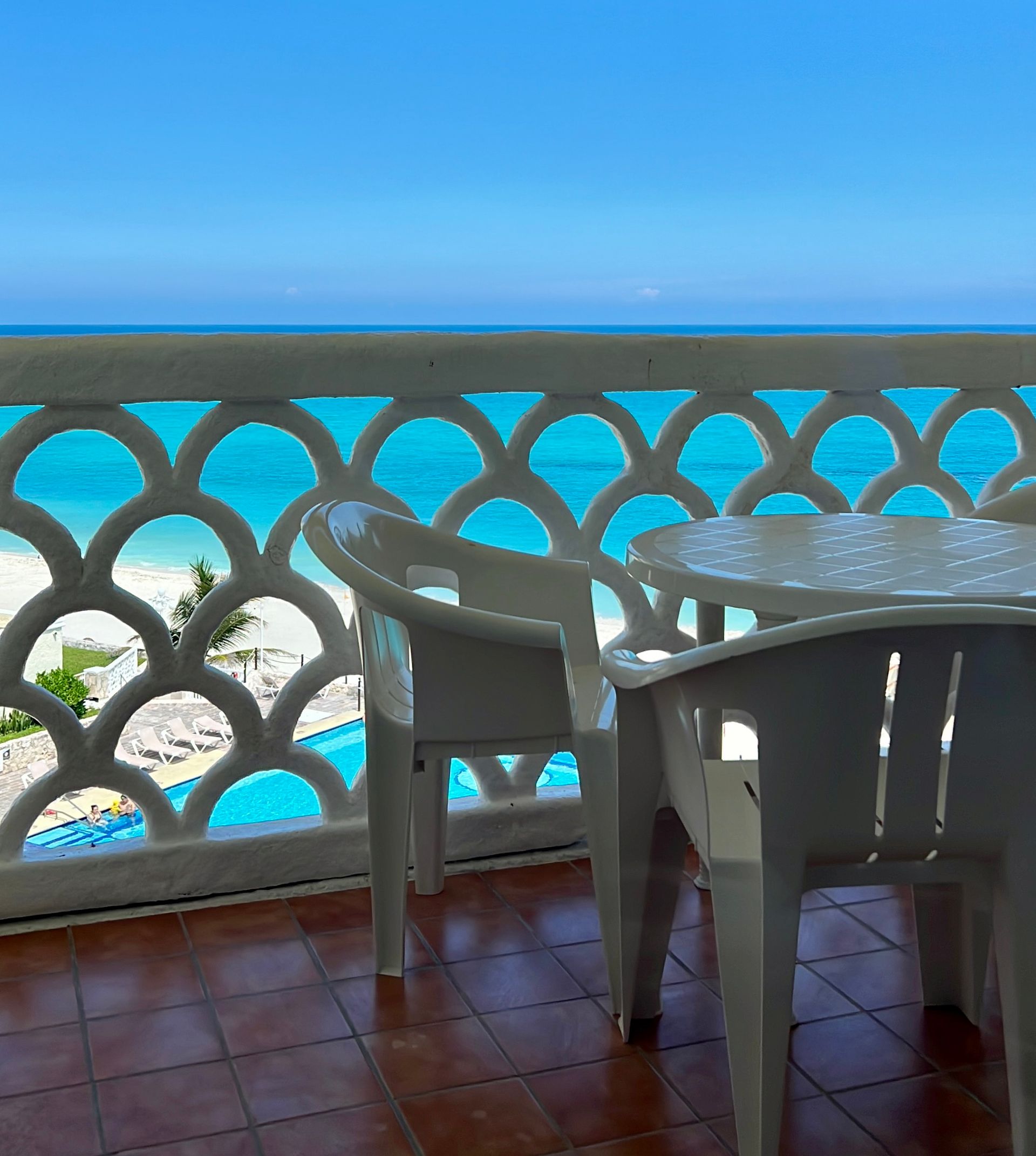 Balcony overlooking turquoise ocean, white plastic chairs and table on brown tiled floor.