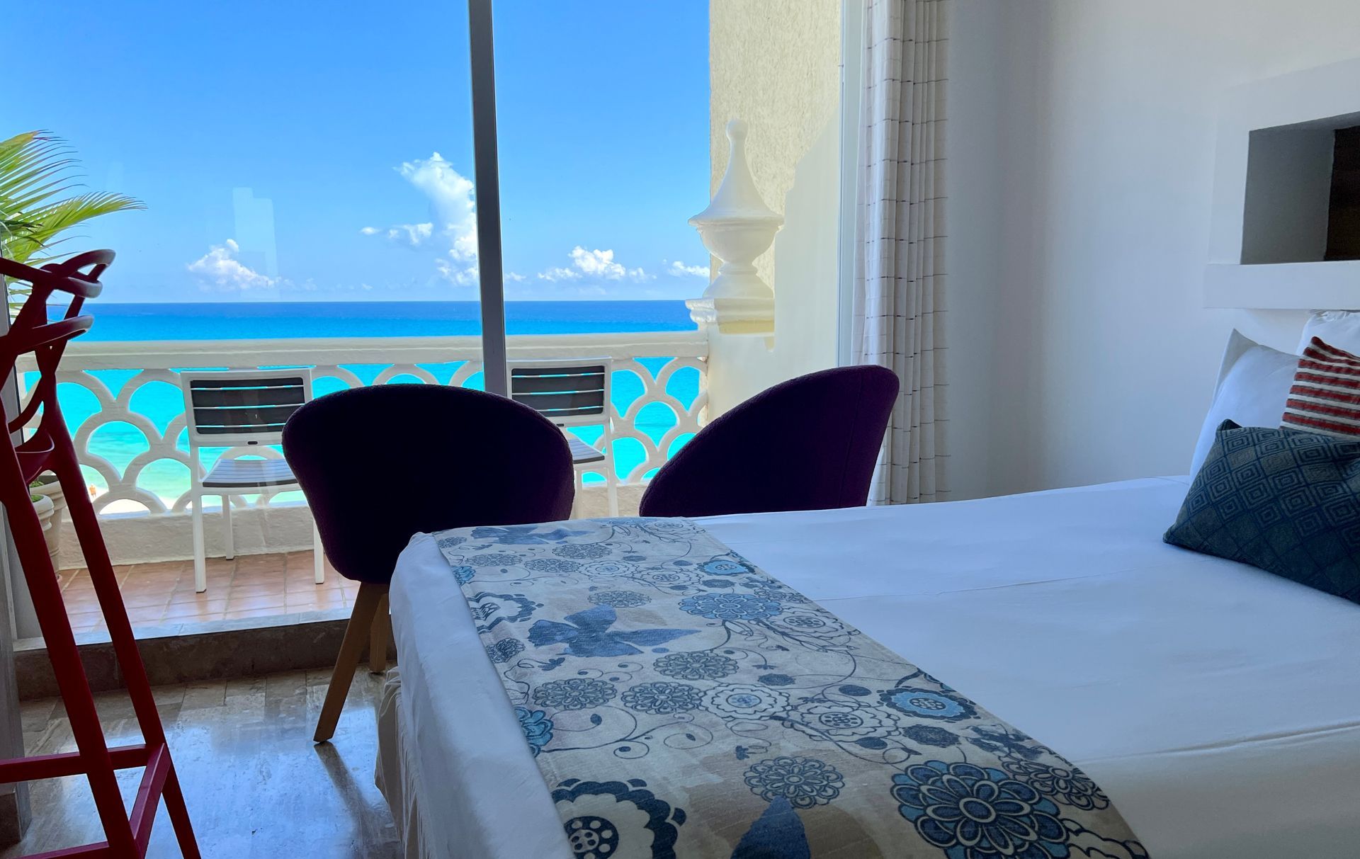 Habitación con vista al mar, cama, sillas y balcón. El océano es azul y el cielo está despejado.