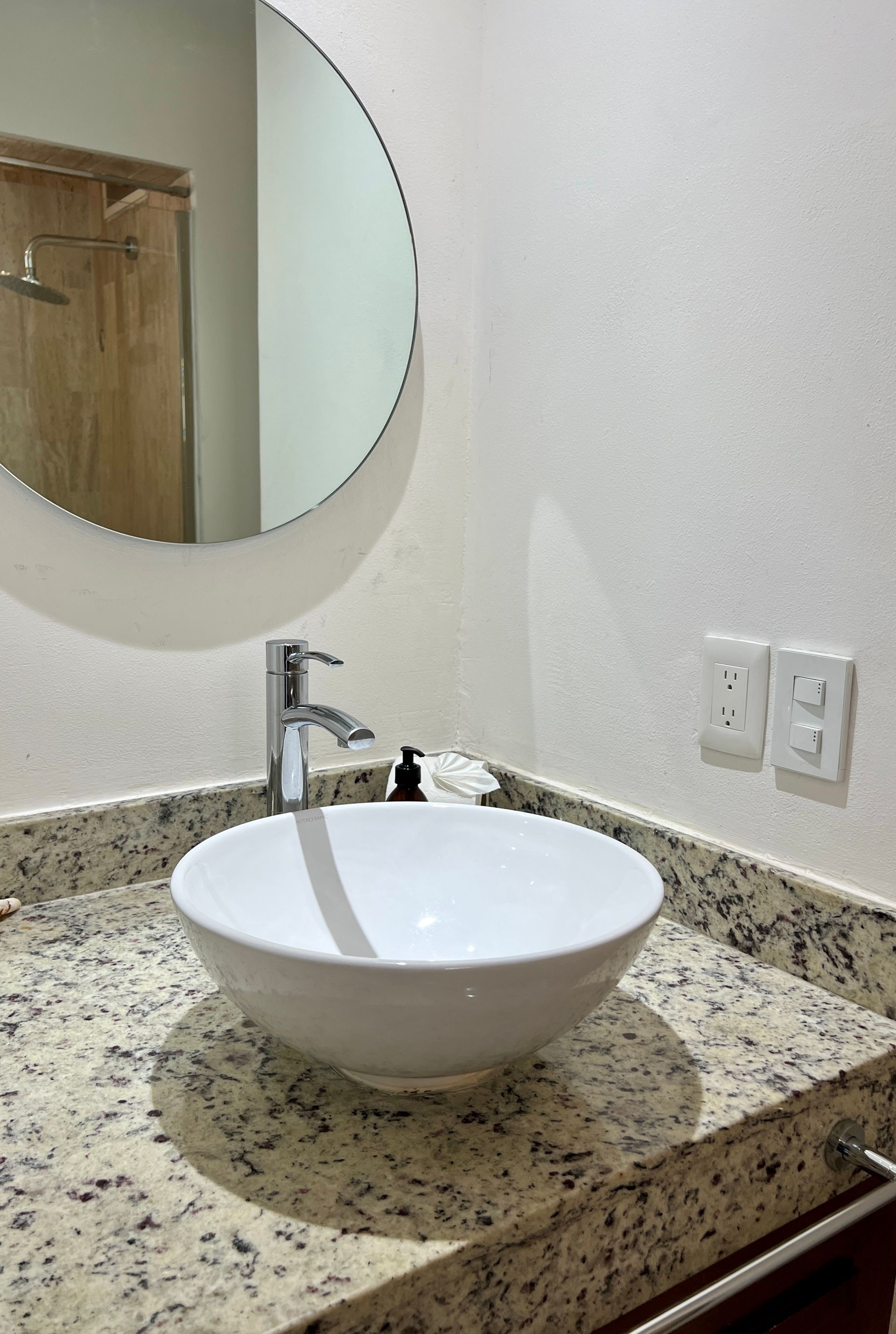 Lavabo blanco sobre encimera de granito con espejo redondo.