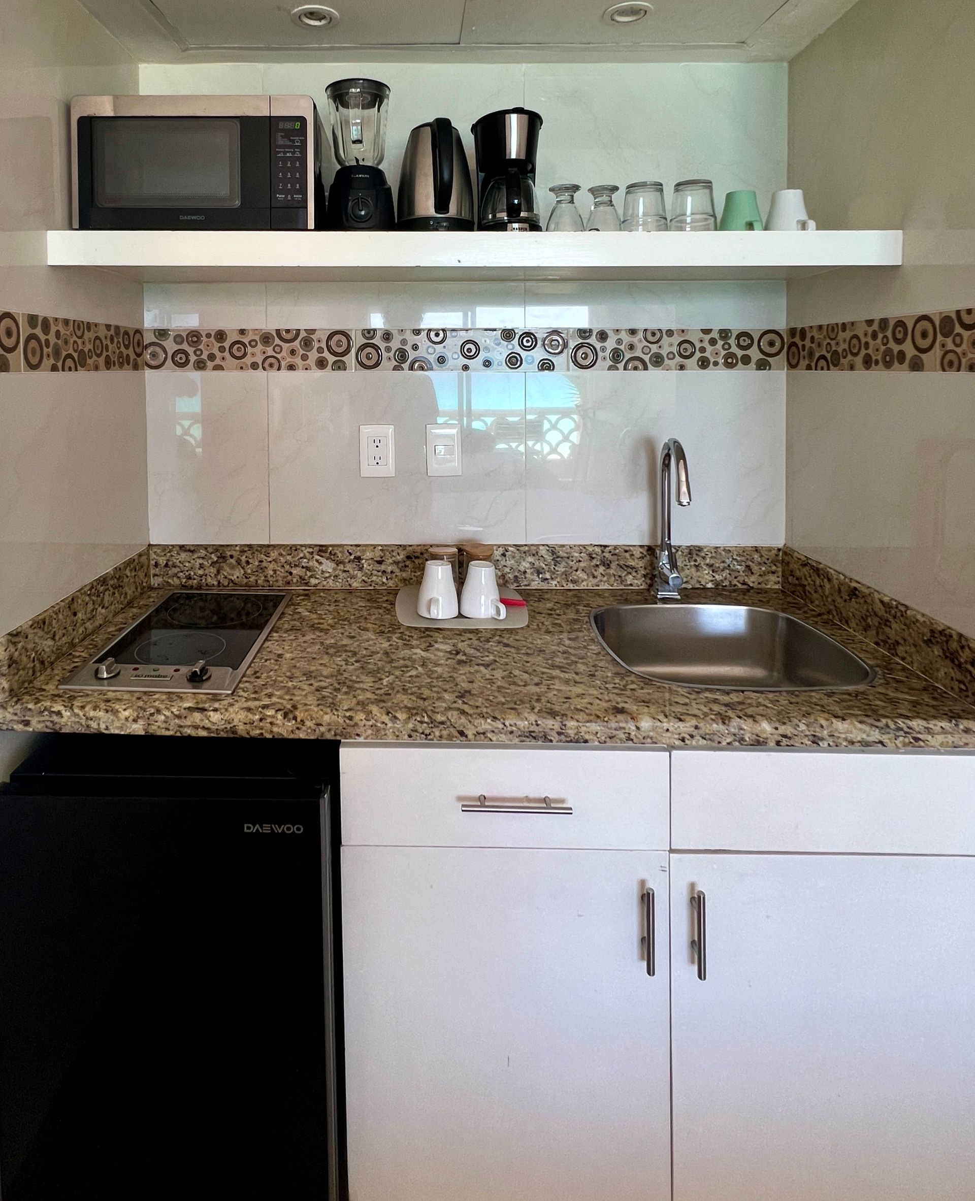 Pequeña cocina con microondas, licuadora, cafetera, fregadero y mini-nevera; gabinetes blancos y encimera de granito.