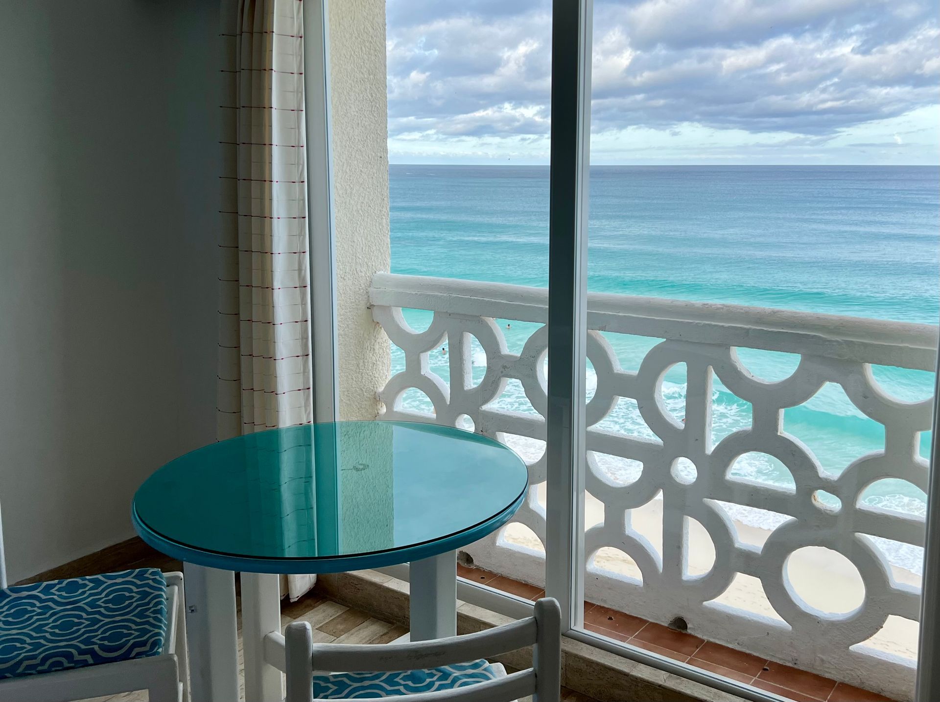 Vista al mar desde el balcón, mesa y sillas turquesas, barandilla decorativa blanca, cielo nublado.