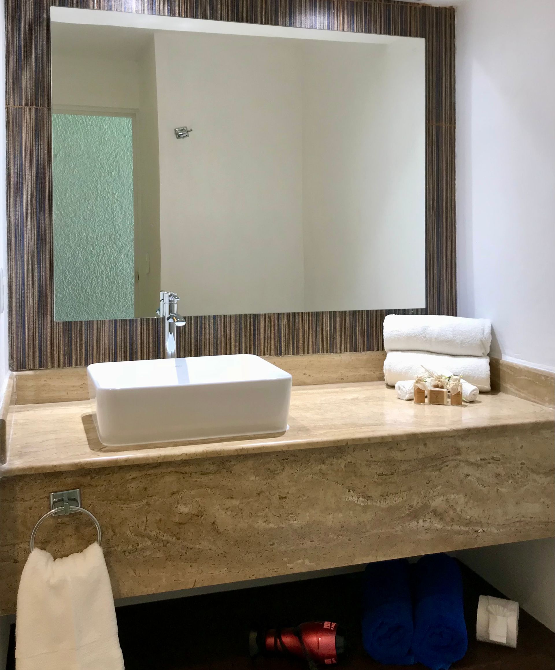 Baño con lavabo rectangular, espejo y toallas. Encimera beige con fondo de azulejos de madera decorativos.