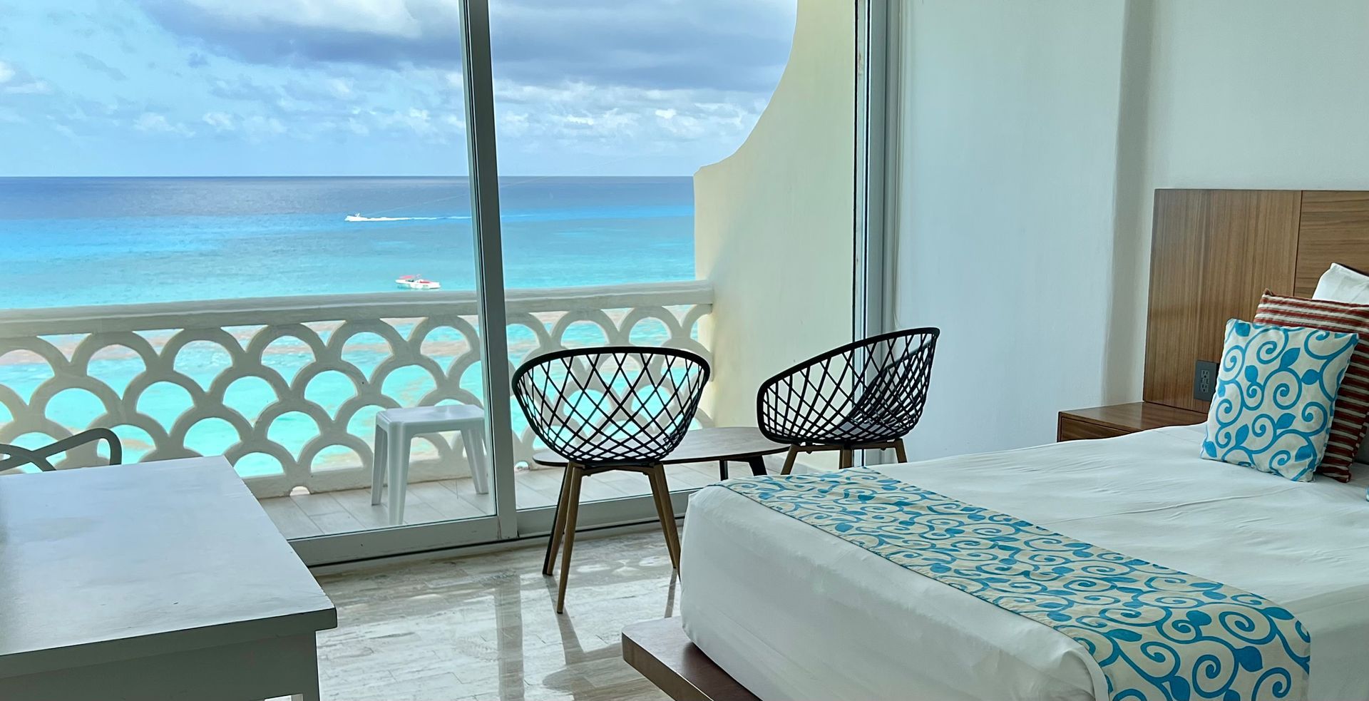Dormitorio con vista al mar, balcón con sillas negras, agua turquesa y cielo.