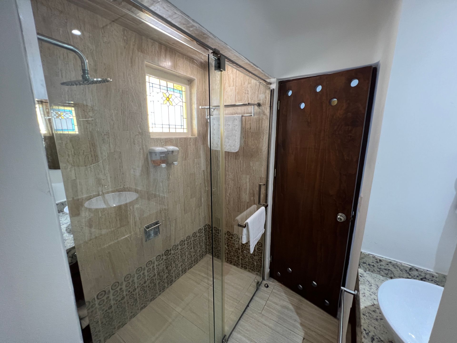 Baño con ducha con mampara de cristal, azulejos beige y puerta de madera oscura con detalles decorativos.