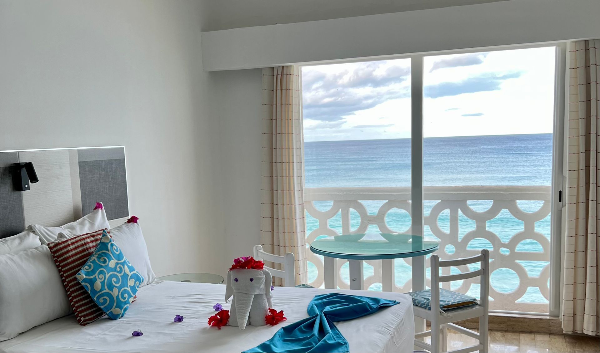 Dormitorio con vista al mar: cama con almohadas, mesa pequeña, agua turquesa, paredes blancas.