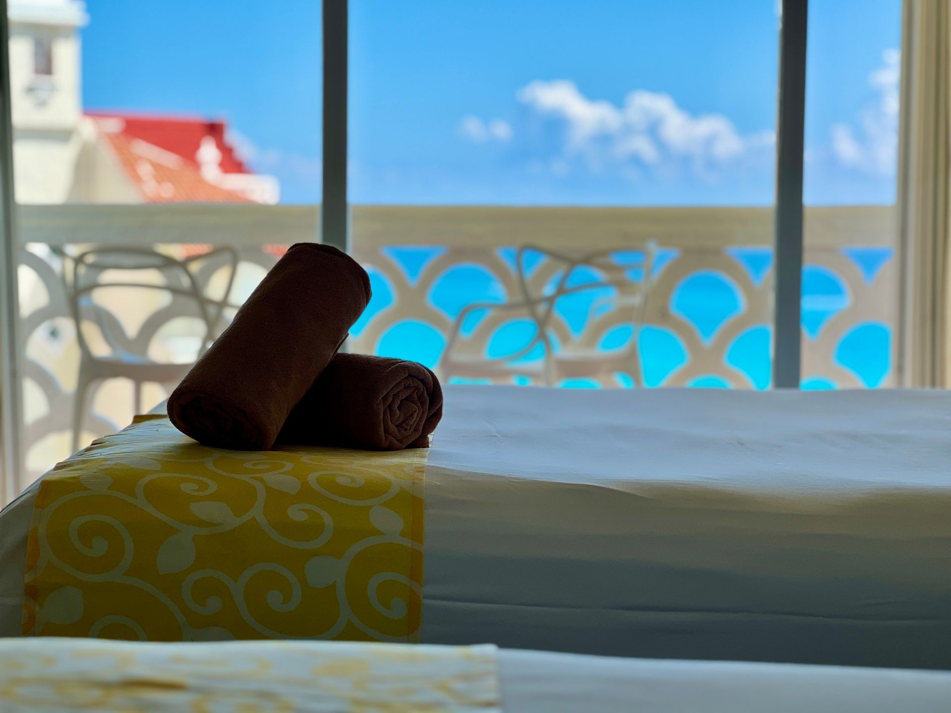 Toallas marrones enrolladas sobre una cama con vista al cielo azul y al edificio desde un balcón.