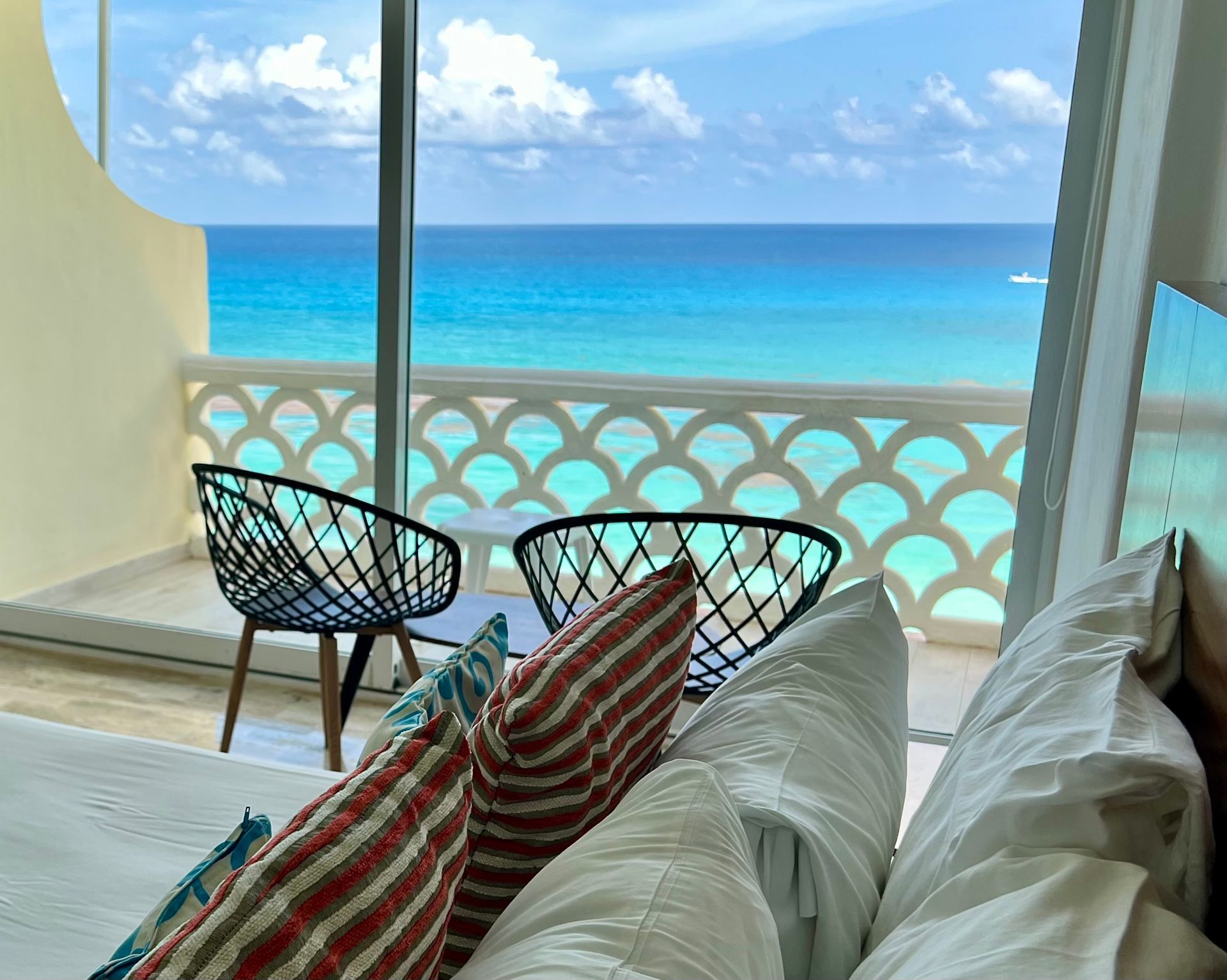 Balcón del dormitorio con vistas al mar turquesa. Dos sillas y mesa, barandilla blanca estampada y cojines coloridos.