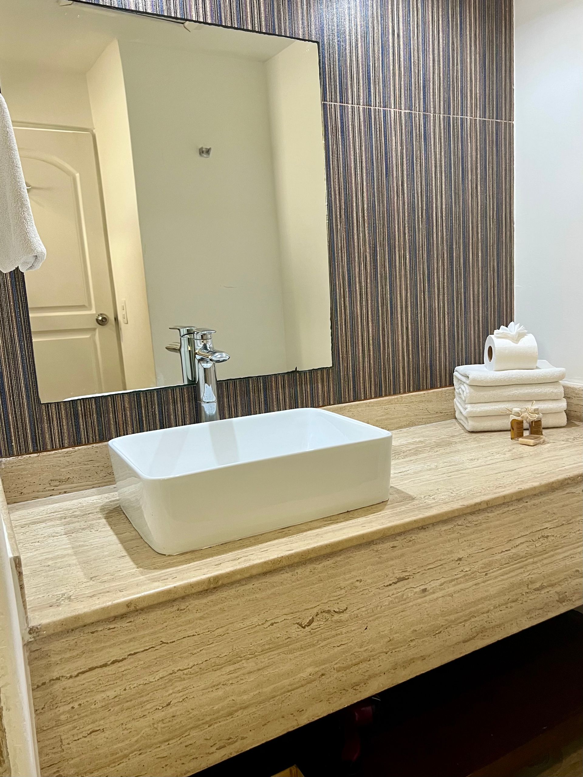 Baño con lavabo rectangular blanco sobre encimera beige, espejo grande y pared marrón texturizada.