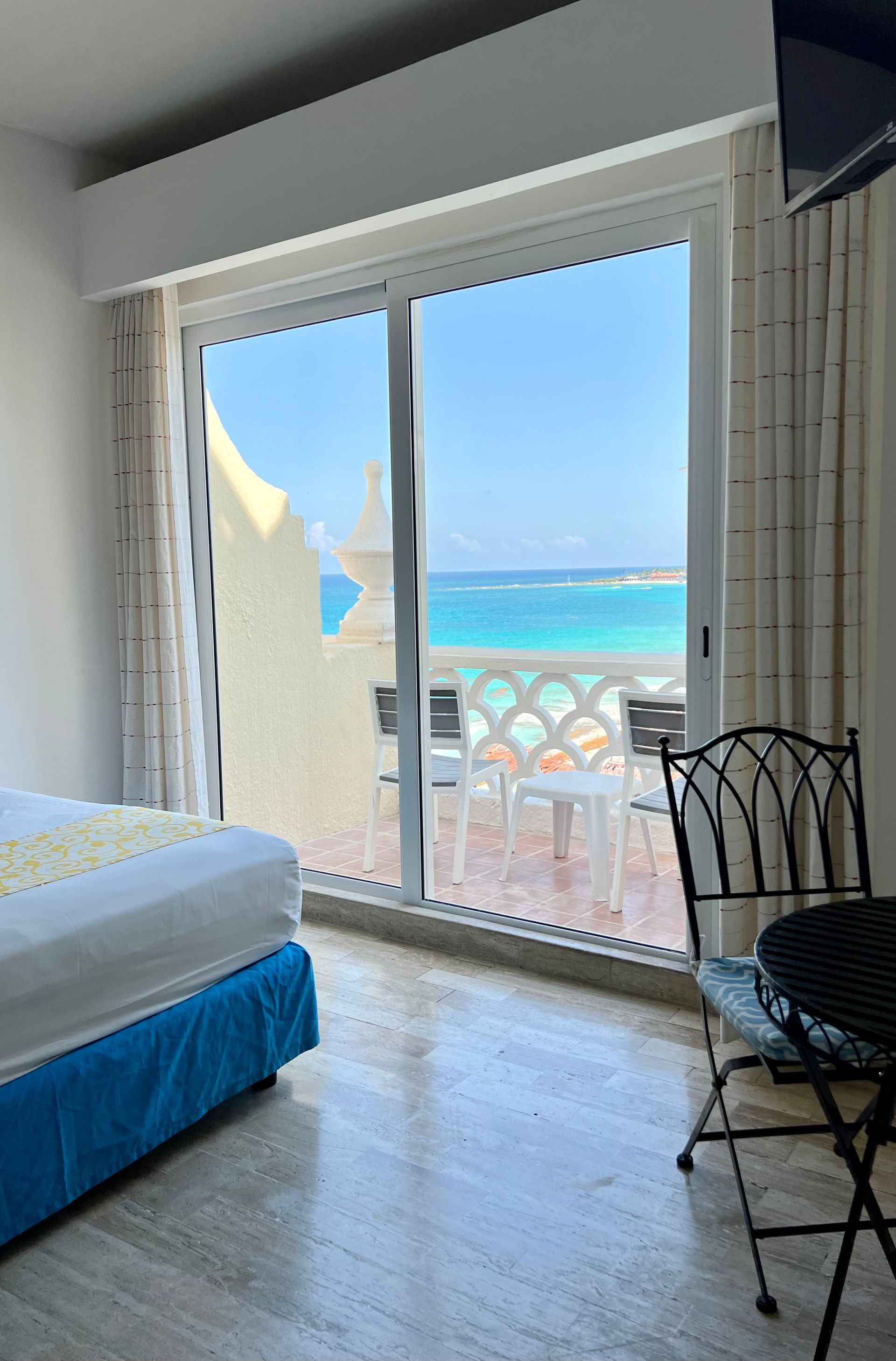 Dormitorio con vista al mar desde el balcón; cama a la izquierda, mesa y silla a la derecha. Agua y cielo azul brillante.