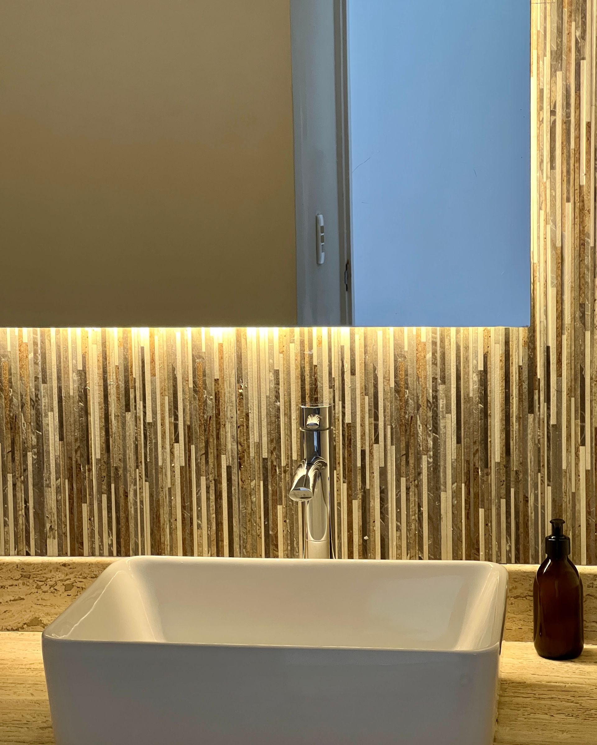 Baño moderno con lavabo rectangular, espejo y pared de mosaico. Dispensador de jabón ámbar sobre encimera.