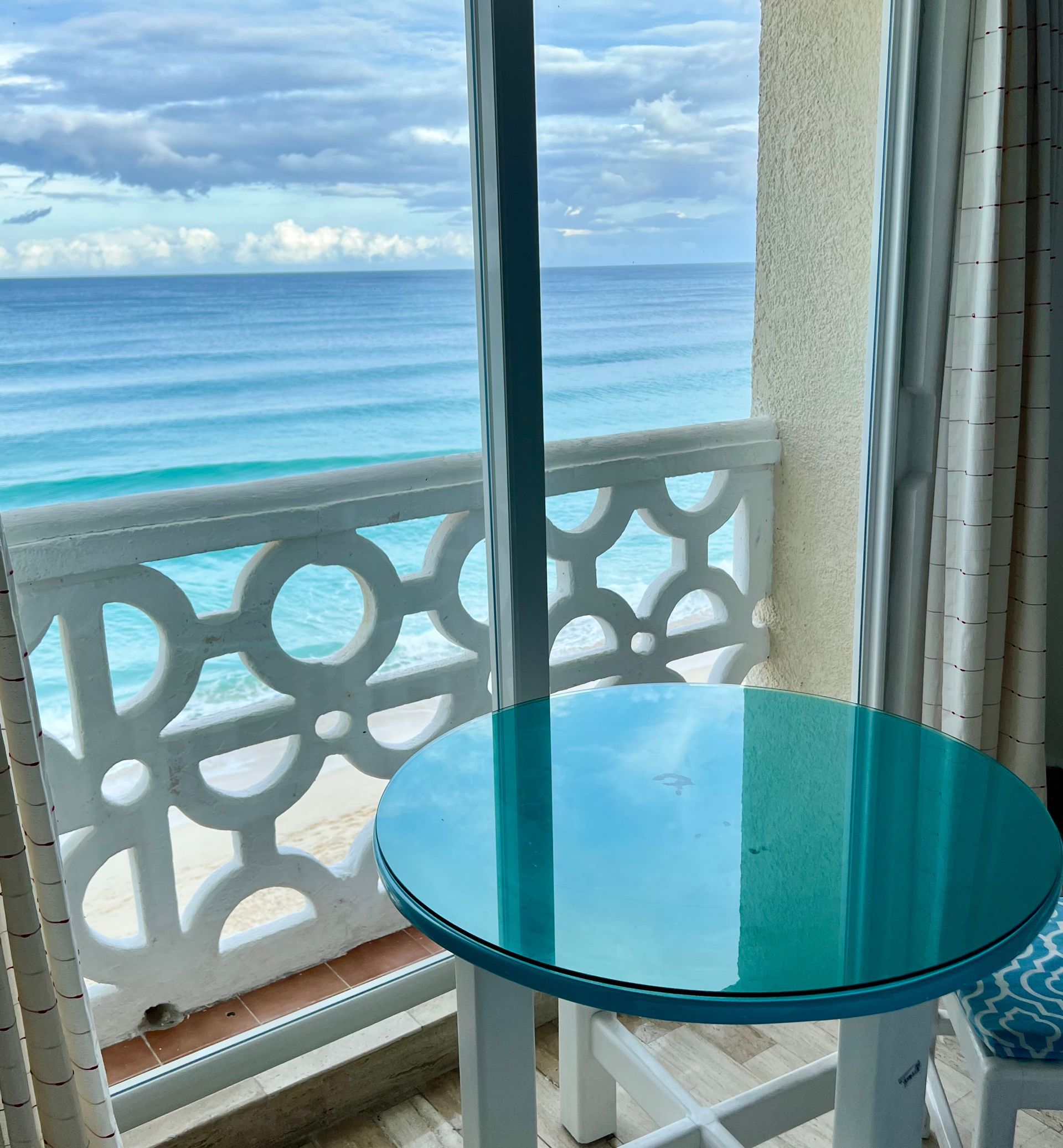Vista al mar desde un balcón, mesa turquesa en primer plano, barandilla blanca, agua azul, cielo nublado.