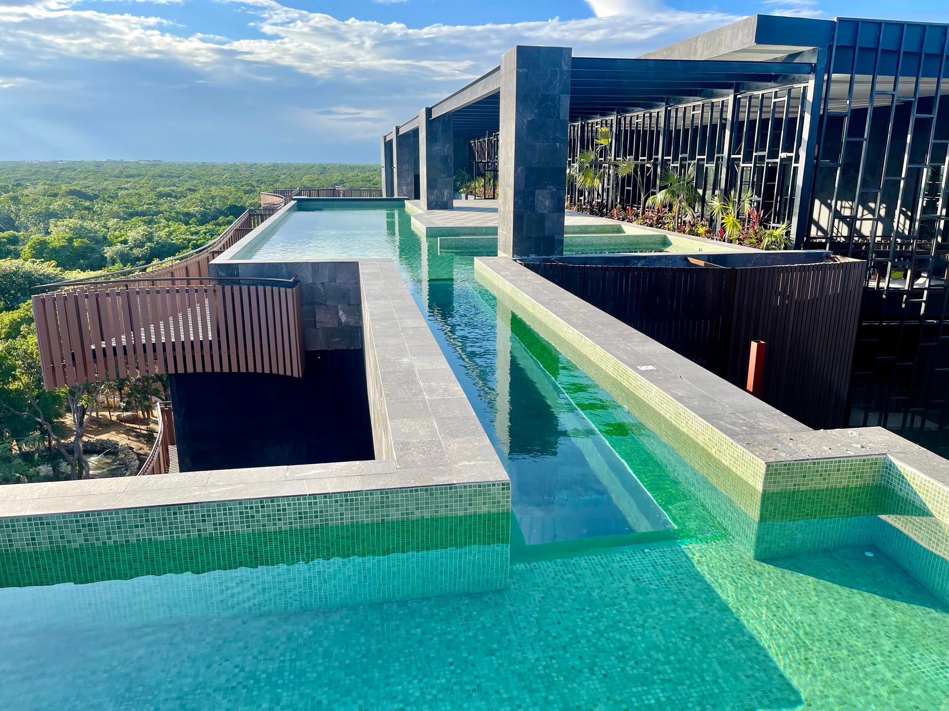 Piscina en la azotea con múltiples secciones, exuberante bosque verde visible al fondo bajo un cielo parcialmente nublado.