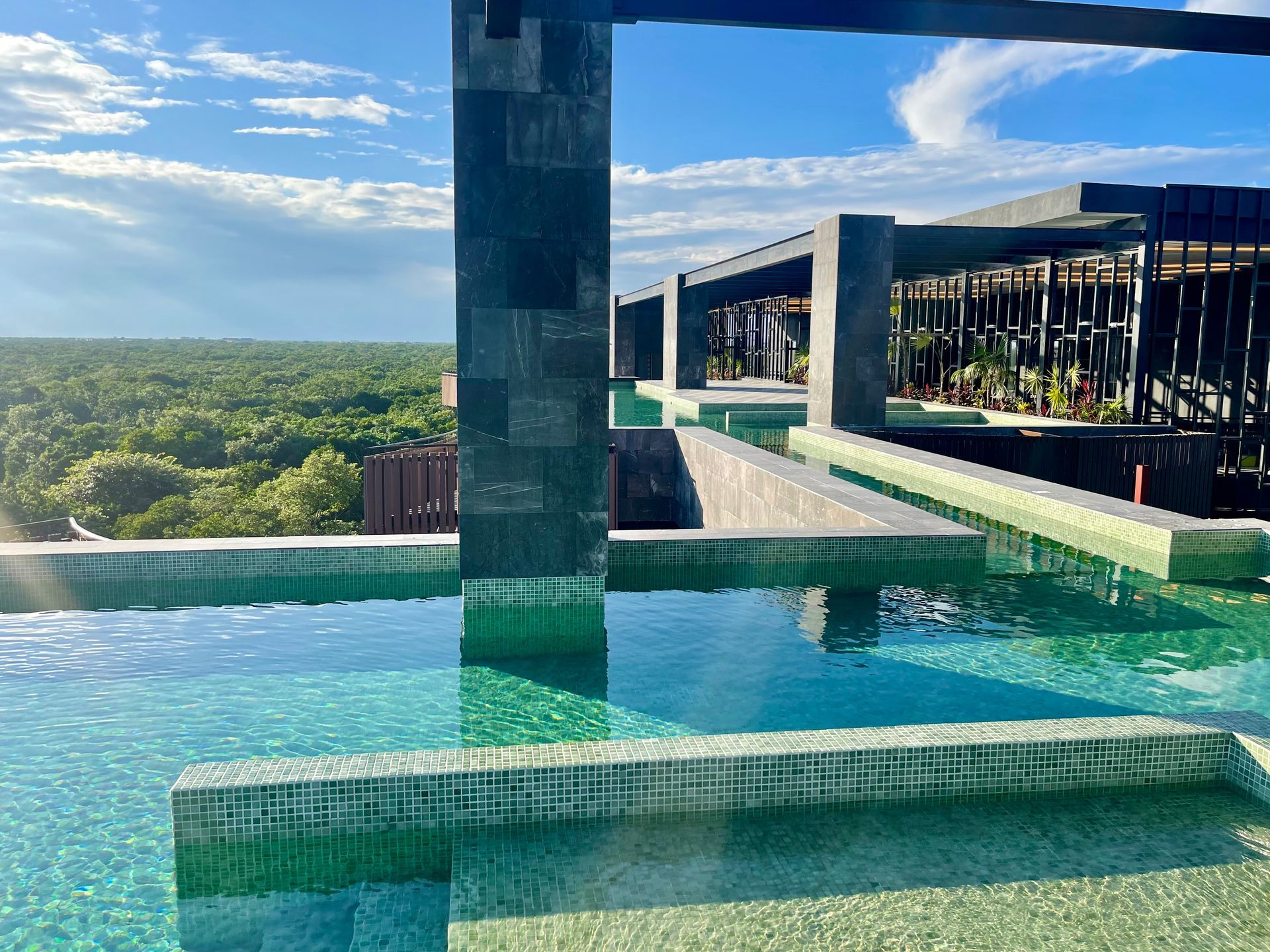 Piscina en la azotea con exuberante vegetación y cielo azul. Arquitectura moderna con agua y columnas.