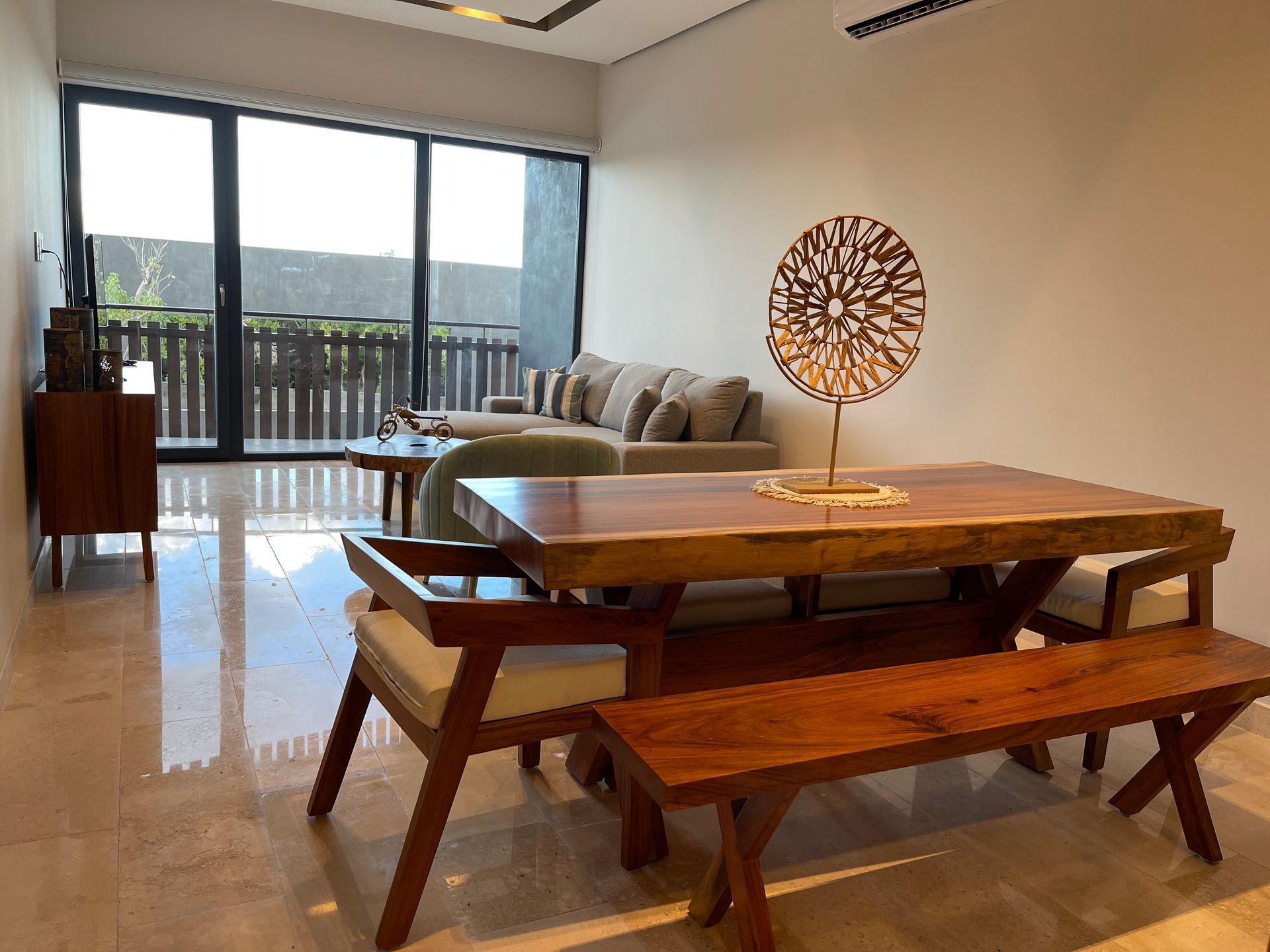 Sala de estar con mesa de comedor de madera, banco y sillas, frente a un sofá y balcón.