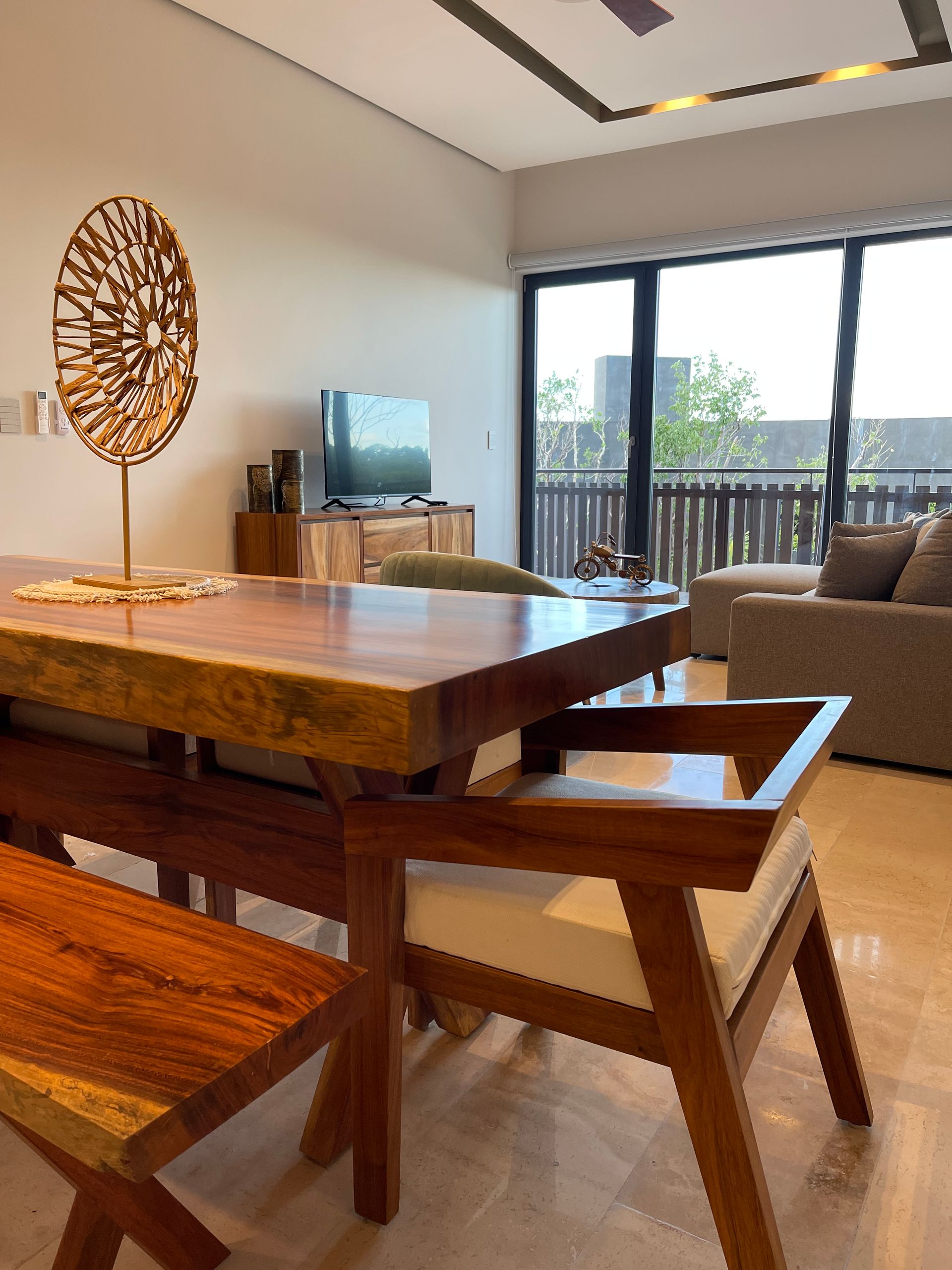 Comedor con mesa de madera, banco y sillas. Se ven un televisor y un sofá.
