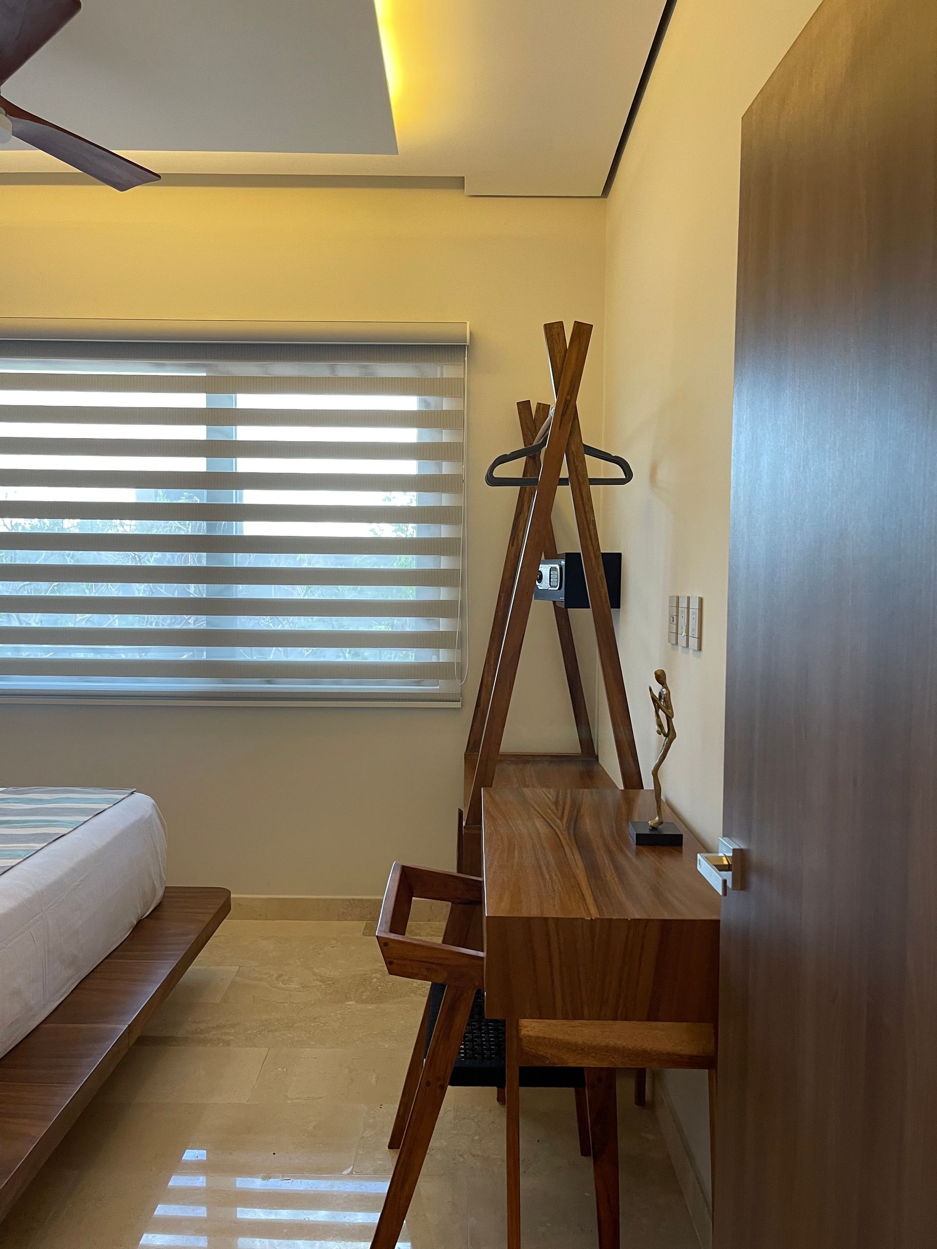 Dormitorio con escritorio de madera, silla, perchero, ventana con persianas y puerta marrón.